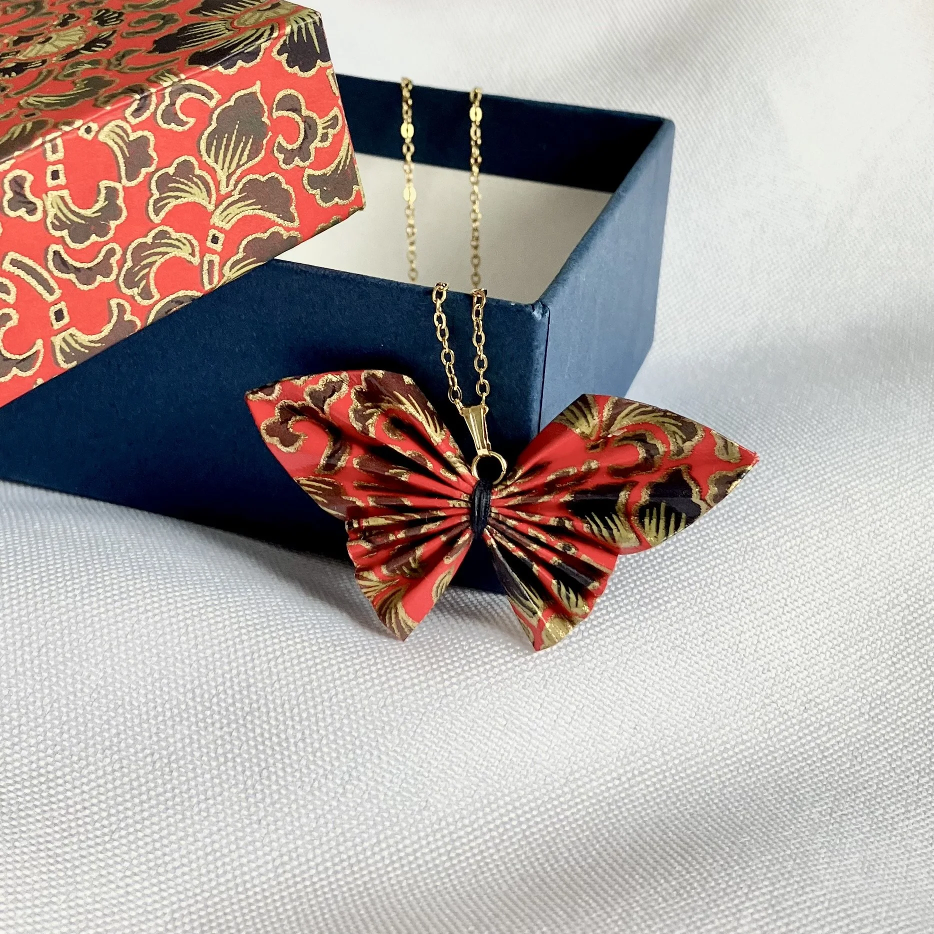 Une boîte décorative rouge et bleue ouverte contenant un collier en forme de papillon rouge en papier japonais avec une chaine doré.