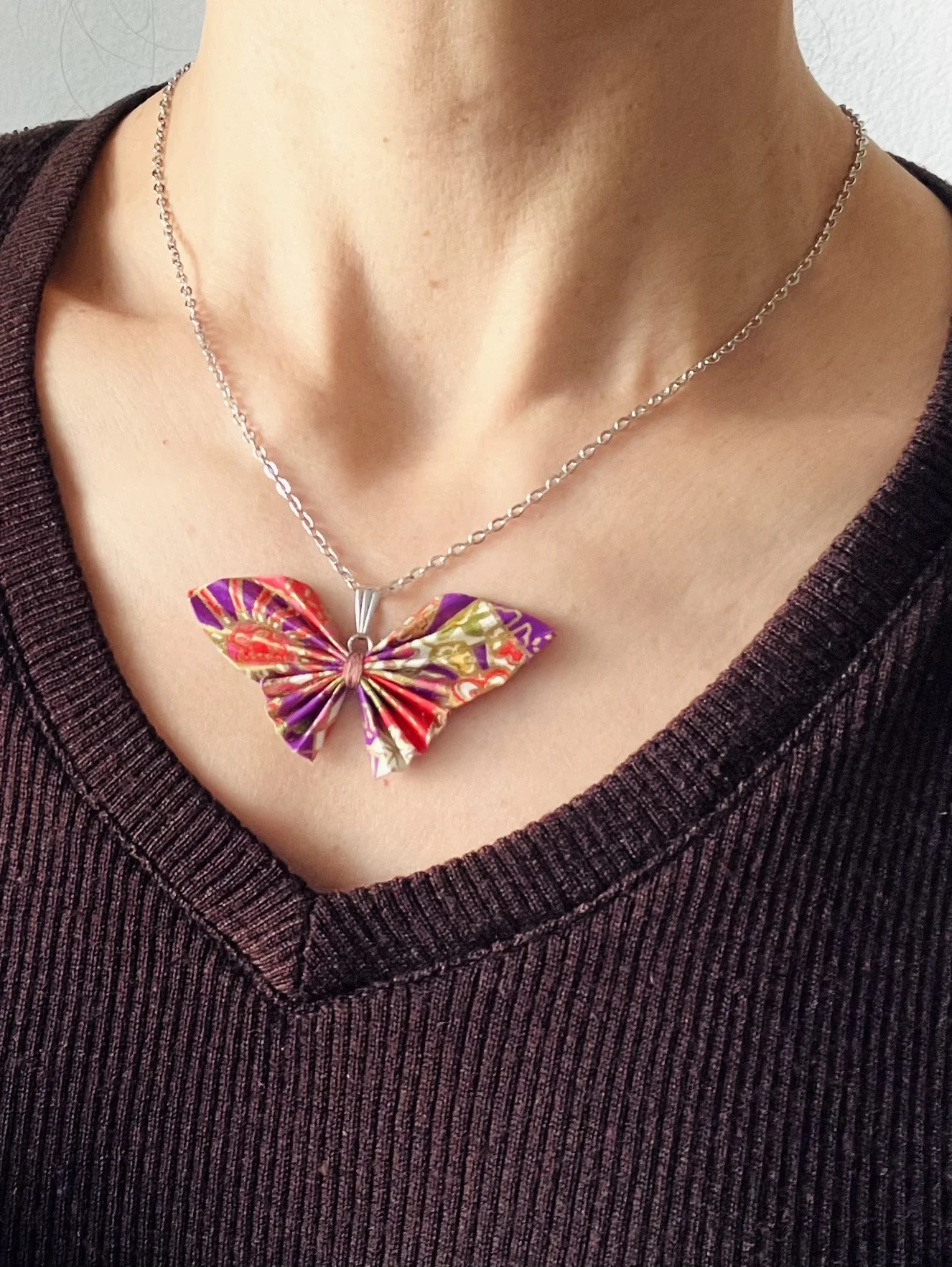 Collier avec pendentif en forme de papillon en billets de papier multicolores, porté par une personne vêtue d'un top en tricot violet foncé.