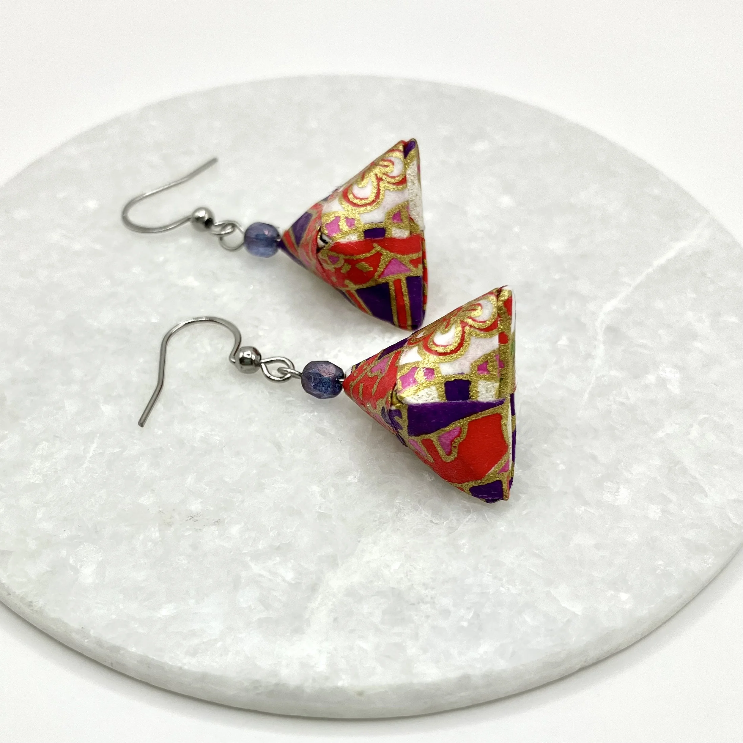 Boucles d'oreilles en papier japonais coloré avec perles en verre violet et crochet en métal acier, posées sur une surface blanche.