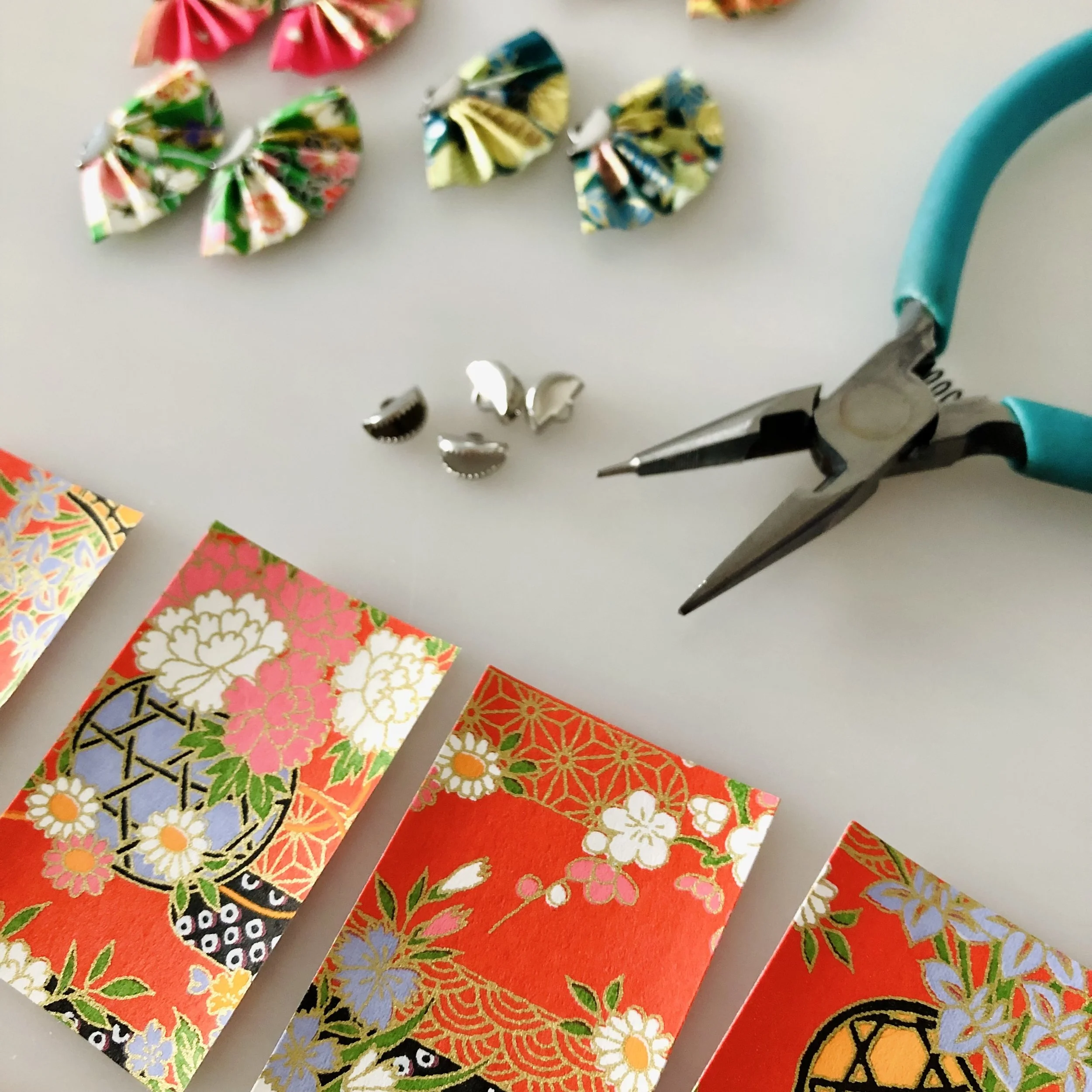 Papier Origami coloré avec motifs floraux et géométriques, disposition en rangées. Présence d'outils de pliage et de découpe.