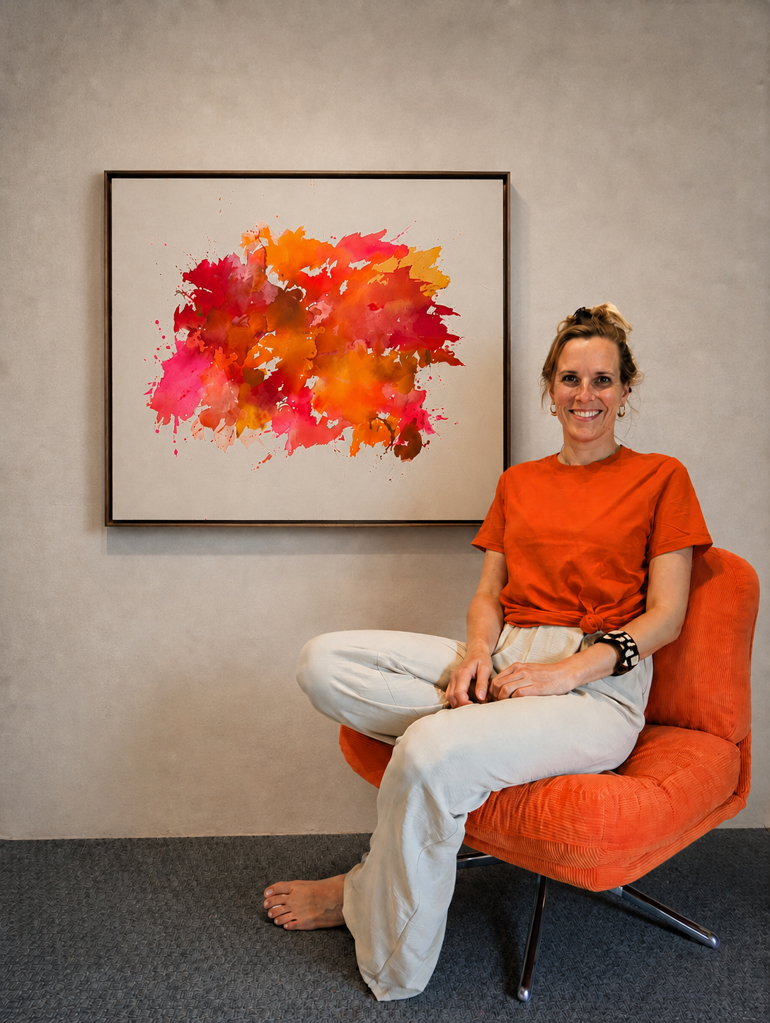 Une femme assise sur un fauteuil orange devant une peinture abstraite aux couleurs orange, rouge, rose et marron.