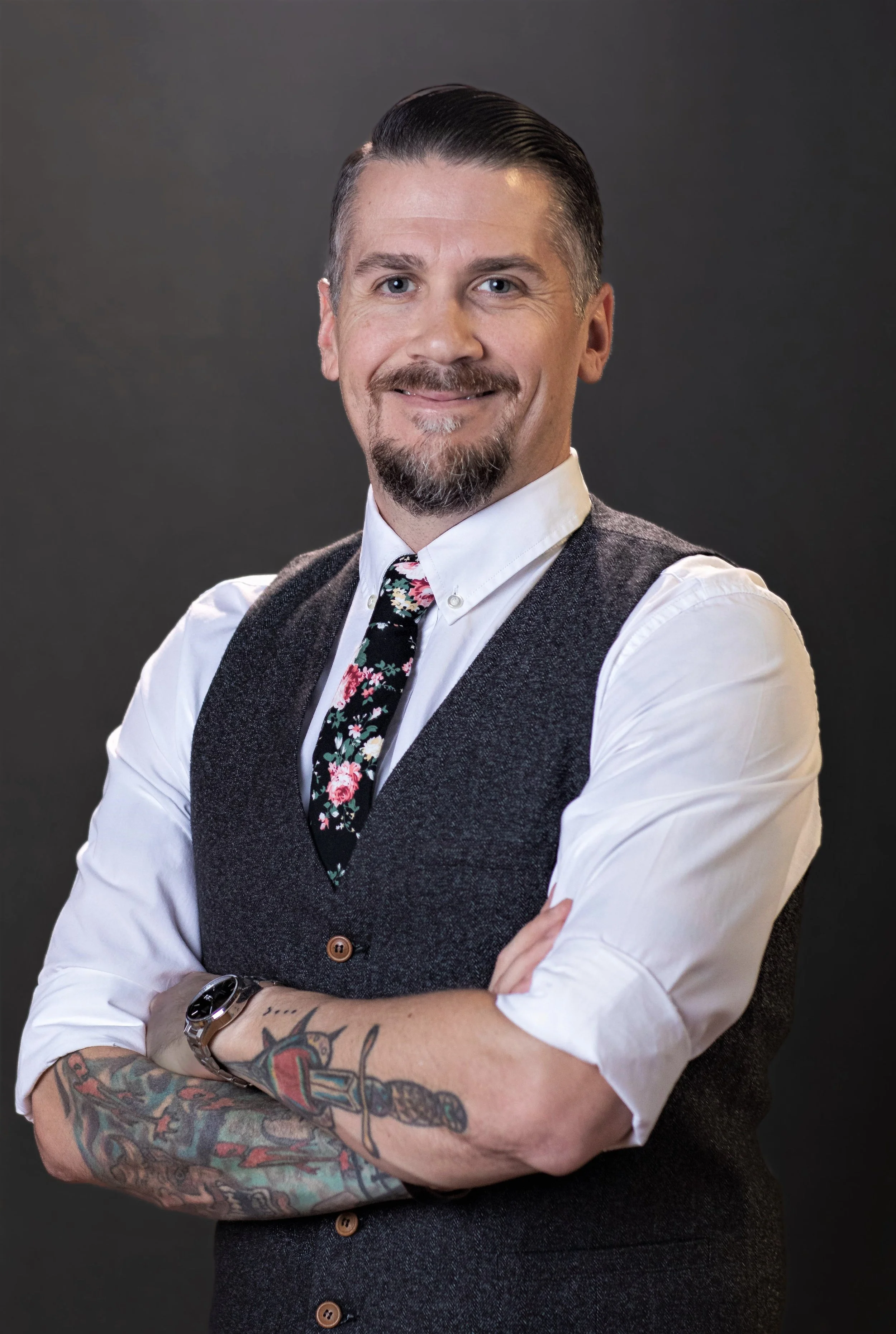 A man with combed-back hair, a goatee, and a floral tie, smiling with arms crossed, wearing a vest, white shirt, and a tattooed left arm, against a dark background.