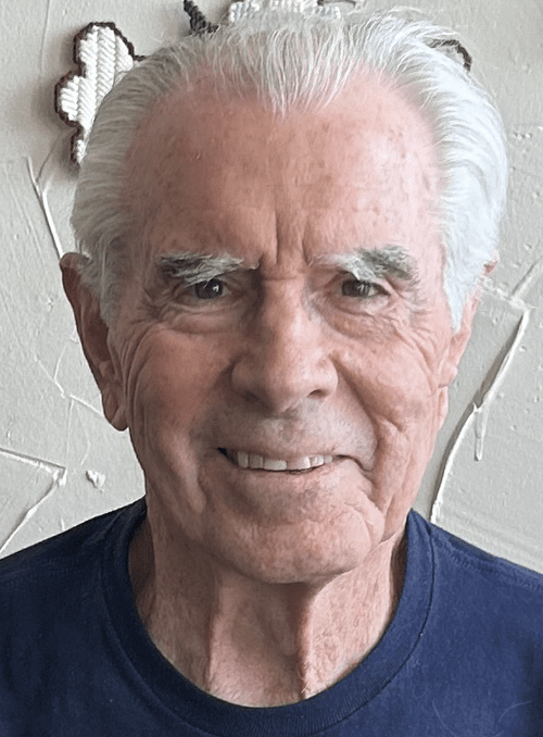 A smiling elderly man with white hair, wearing a dark blue shirt, standing in front of a textured wall with decorative elements.