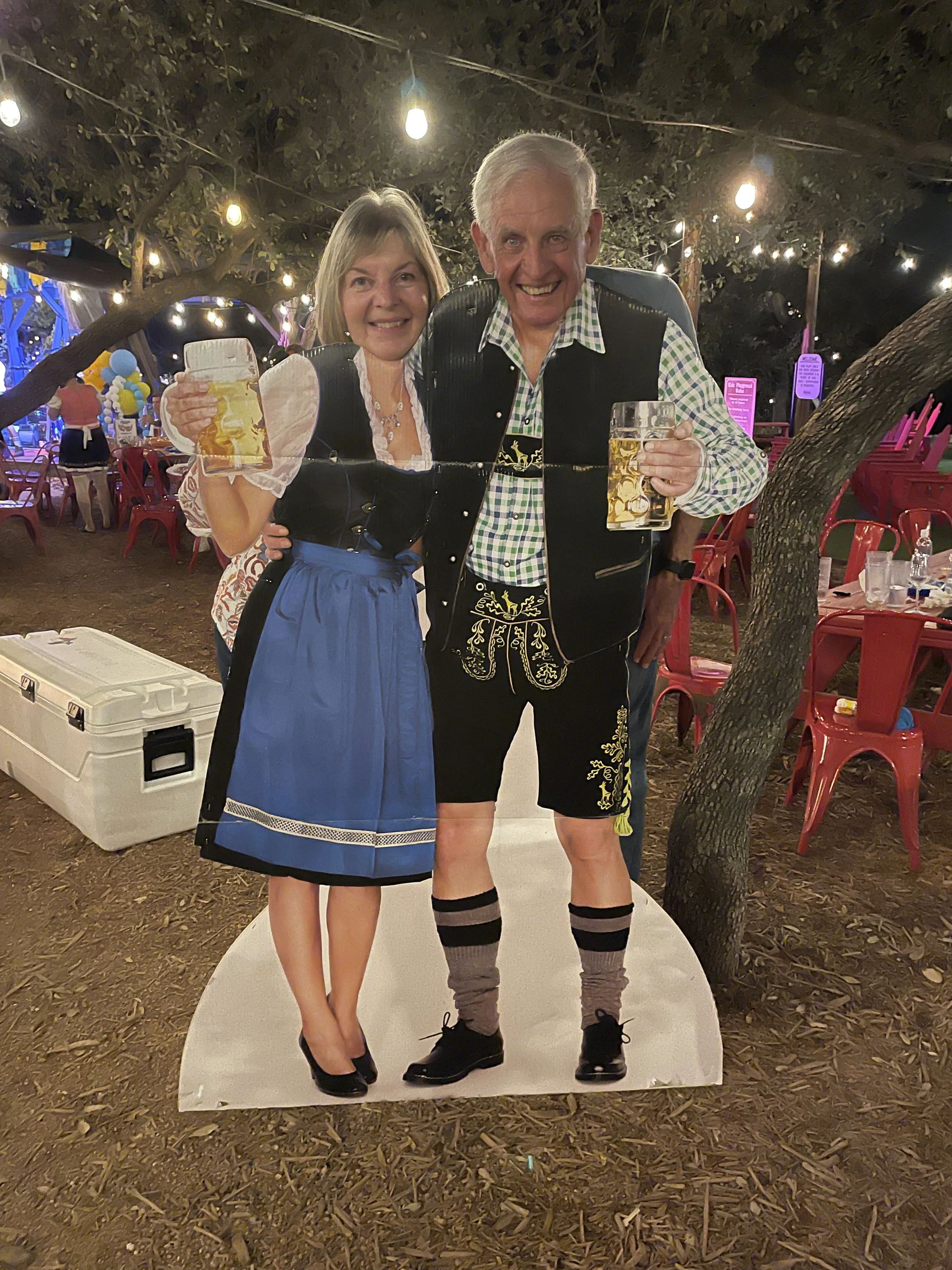 Two people, a woman and a man, standing on a cutout of a couple holding beer mugs at an outdoor event during nighttime, with string lights above and tables with red chairs in the background.