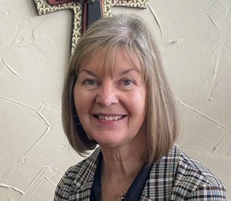 A smiling woman with shoulder-length gray hair, wearing a patterned blouse, standing in front of a light-colored wall with decorative items.