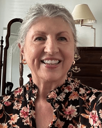 A smiling woman with gray hair, wearing a floral top, standing indoors with a mirror and a dresser in the background.