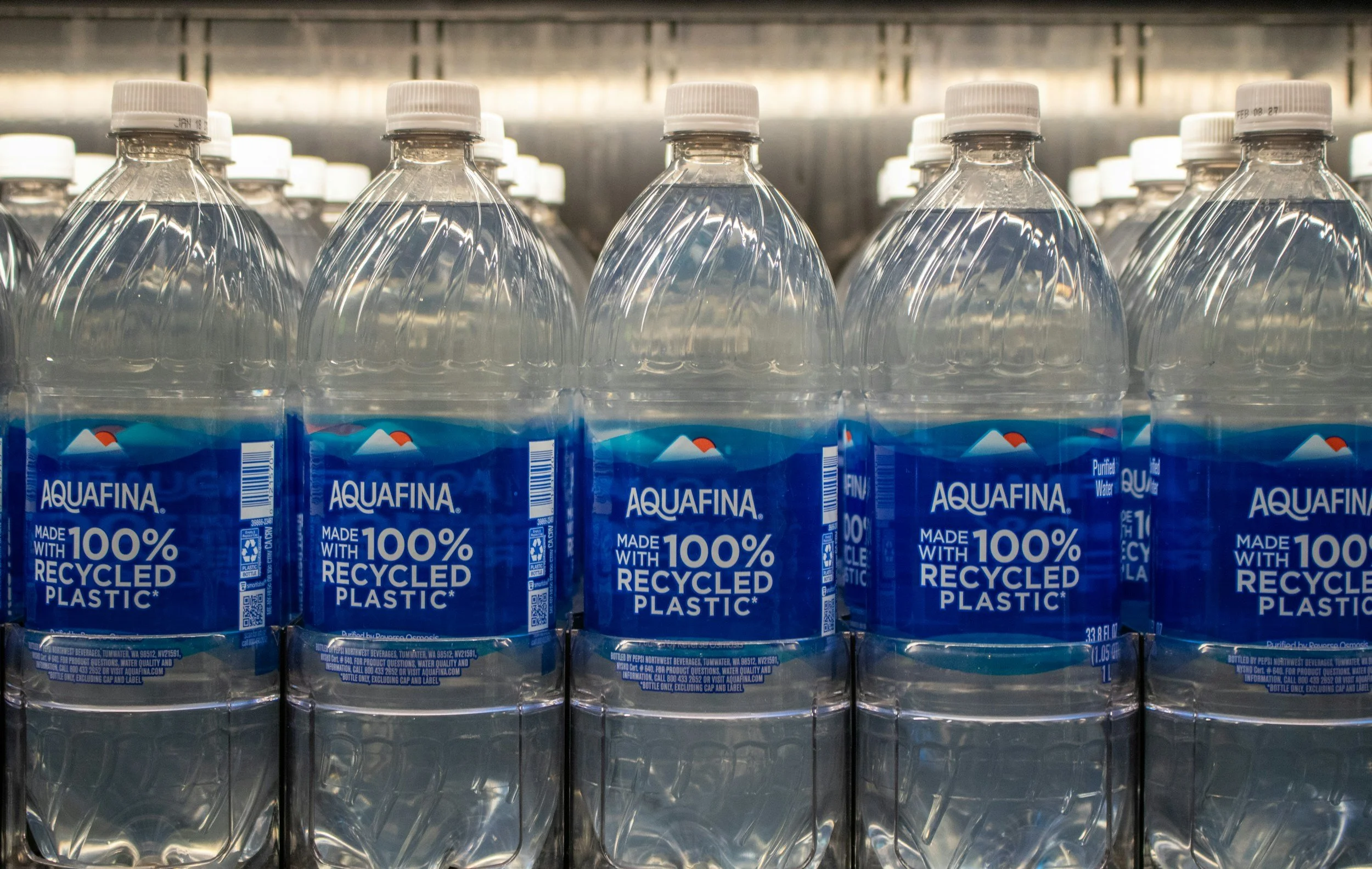 Rows of Aquafina bottled water on store shelves, labeled as made with 100% recycled plastic.
