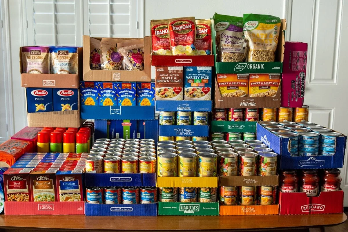 Multiple boxes and cans of food and soup on a table, including pasta, canned soup, and dry goods.