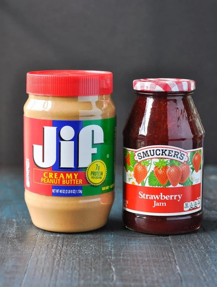 A jar of Jif creamy peanut butter and a jar of Smucker's strawberry jam on a wooden surface against a black background.