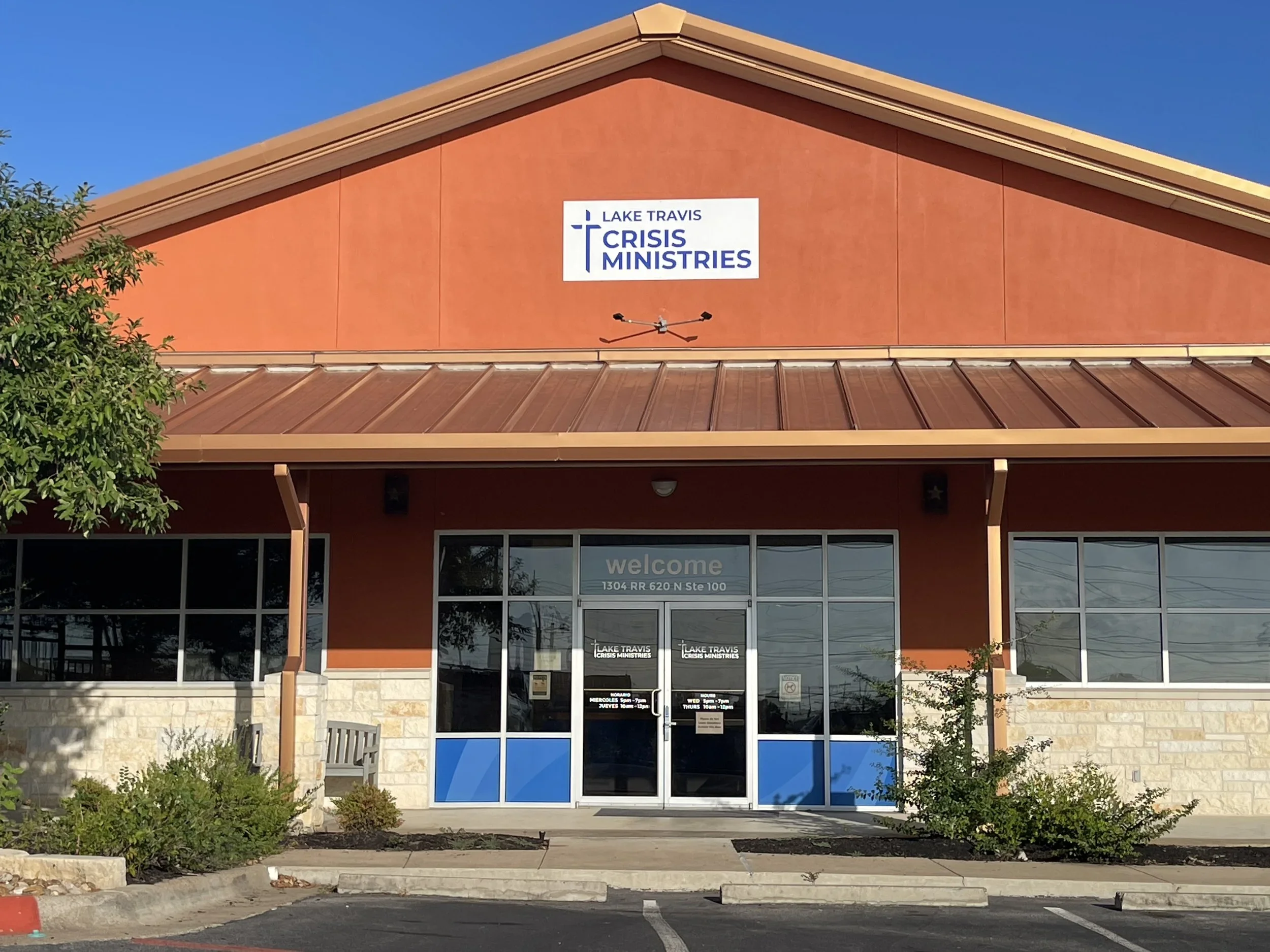 The front view of a building with a sign that reads 'Lake Travis Crisis Ministries' and an entrance door labeled 'welcome', with windows, plants, and a parking lot in front.