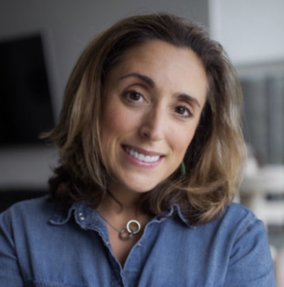 A woman with long brown, wavy hair, wearing eyeglasses on her head, smiling, dressed in a colorful top with yellow and green patterns.