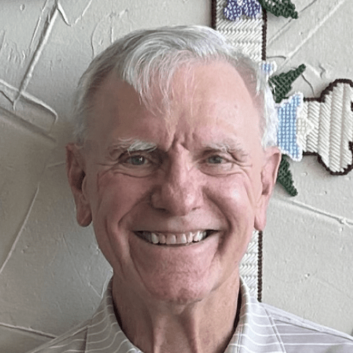 A smiling elderly man with white hair in front of a wall with a cross and flower decoration.