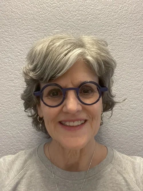 A smiling older woman with gray, wavy hair and blue glasses, wearing a light-colored shirt and earrings, standing against a textured wall.