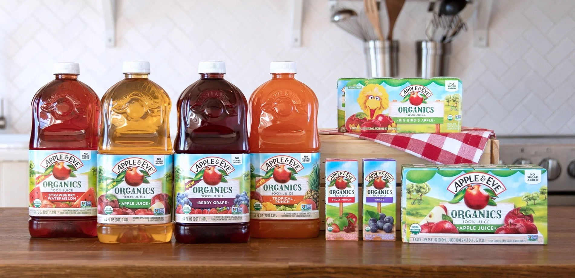 Various bottles and cartons of Apple & Eve organic juices, including strawberry watermelon, apple, berry grape, tropical punch, and grape, arranged on a wooden surface in a kitchen setting.