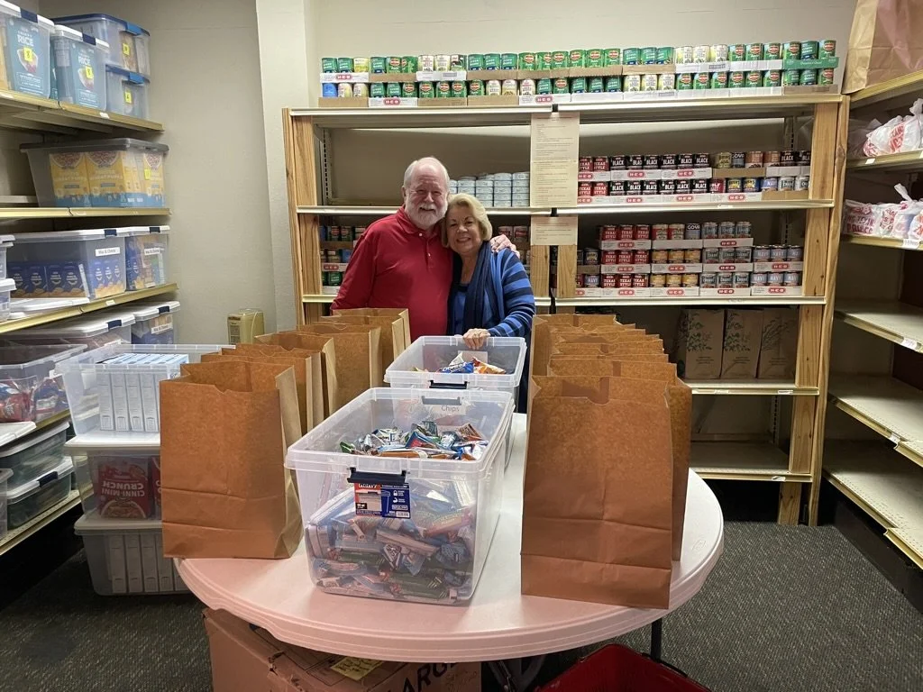 Two seniors smiling and hugging in a food bank supply room with shelves of canned goods and other items behind them, and paper bags and a box of snacks on a round table in front.