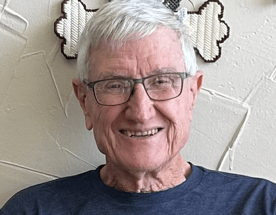 A smiling elderly man with glasses, short white hair, wearing a dark blue shirt, sitting indoors with a decorative wall hanging behind him.