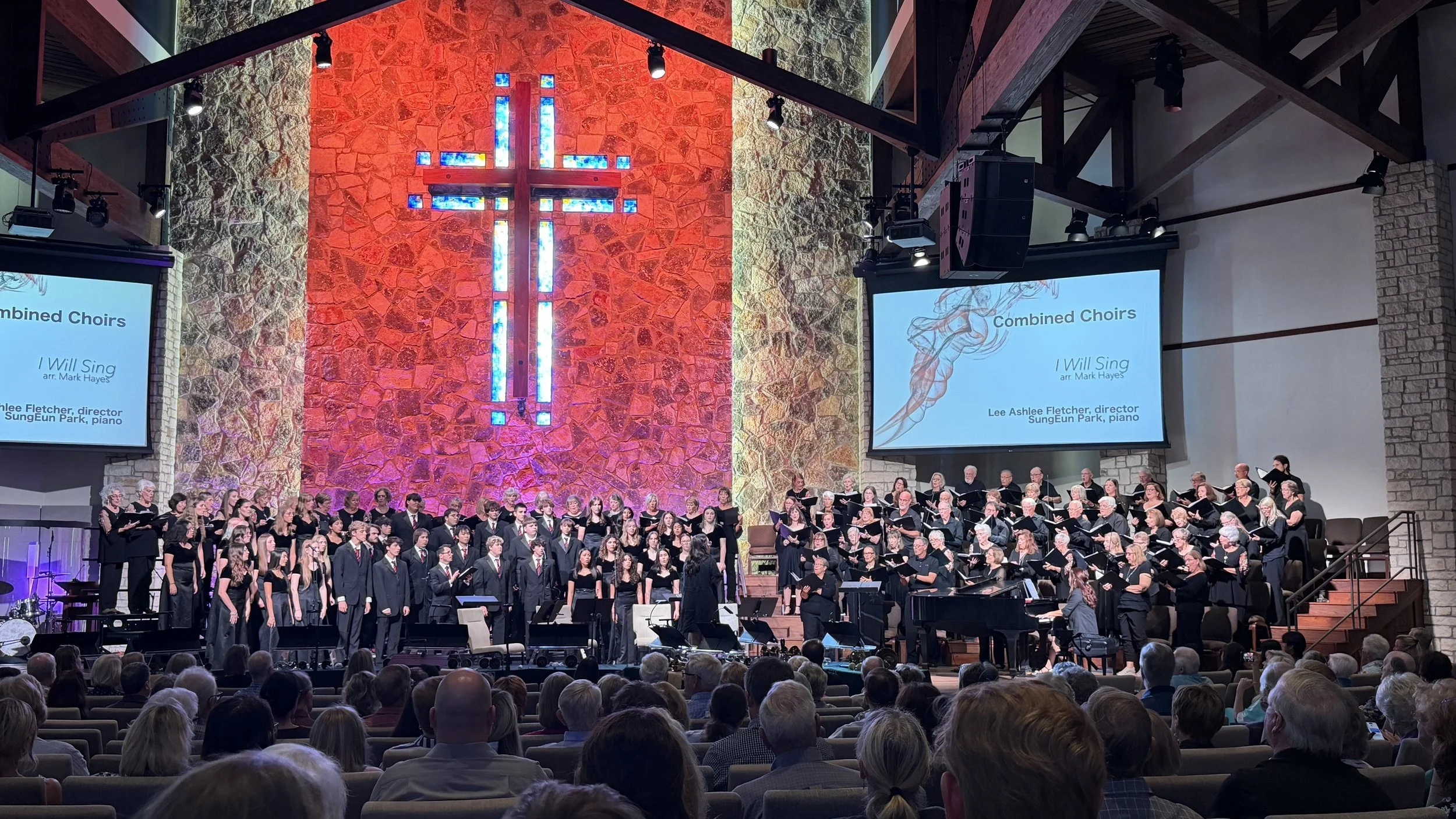 A large choir performing in a church with stained glass cross in the background, and a congregation watching in the pews.