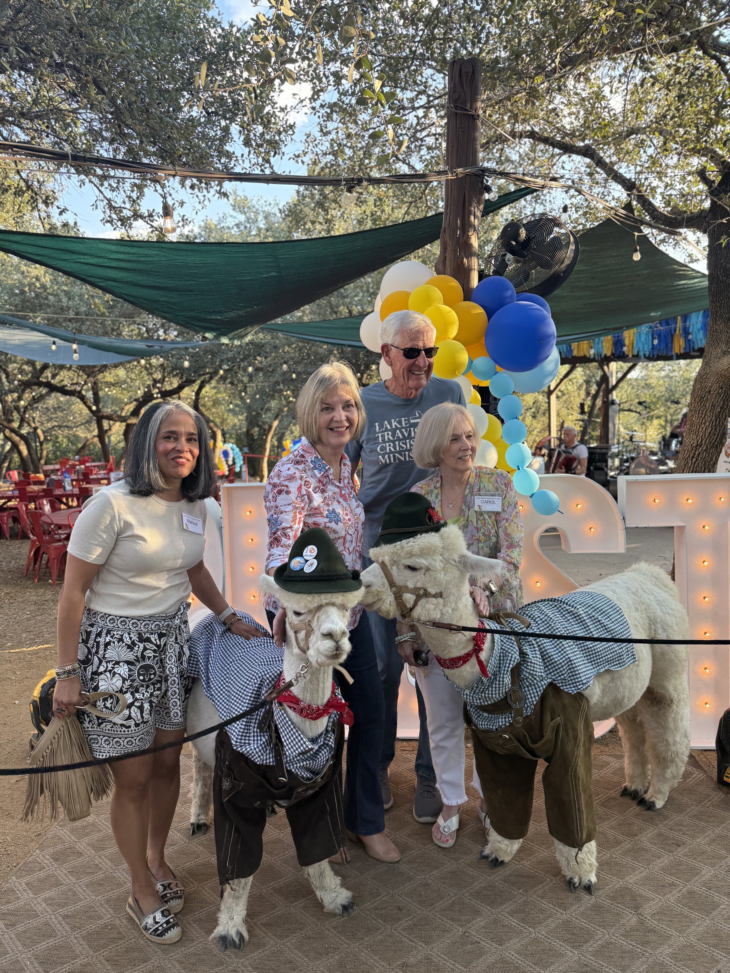 Four women and one man posing with two llamas dressed in traditional German attire at an outdoor festival. The background features colorful balloons, large illuminated letters, and trees with string lights. The scene appears festive and lively.