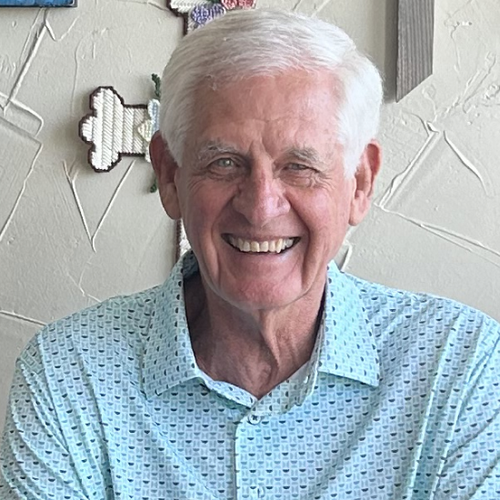 Smiling elderly man with white hair in a light blue patterned shirt, sitting indoors with wall art in the background.