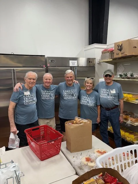 Five volunteers in matching blue T-shirts standing side by side in a kitchen, smiling, with arms around each other. There is a table with food items and a red basket in front of them.