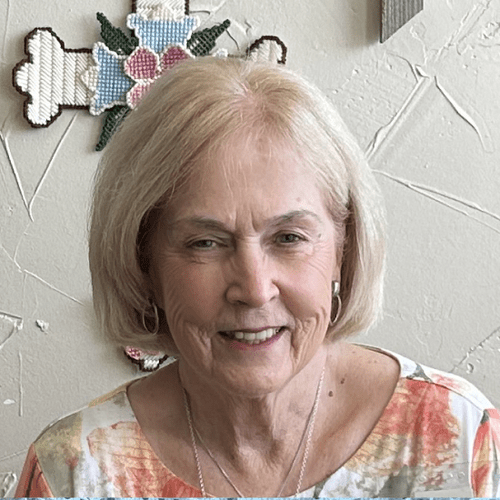 Smiling elderly woman with short blonde hair, wearing a colorful blouse, in front of a textured wall with floral cross-stitch embroidery in the background.