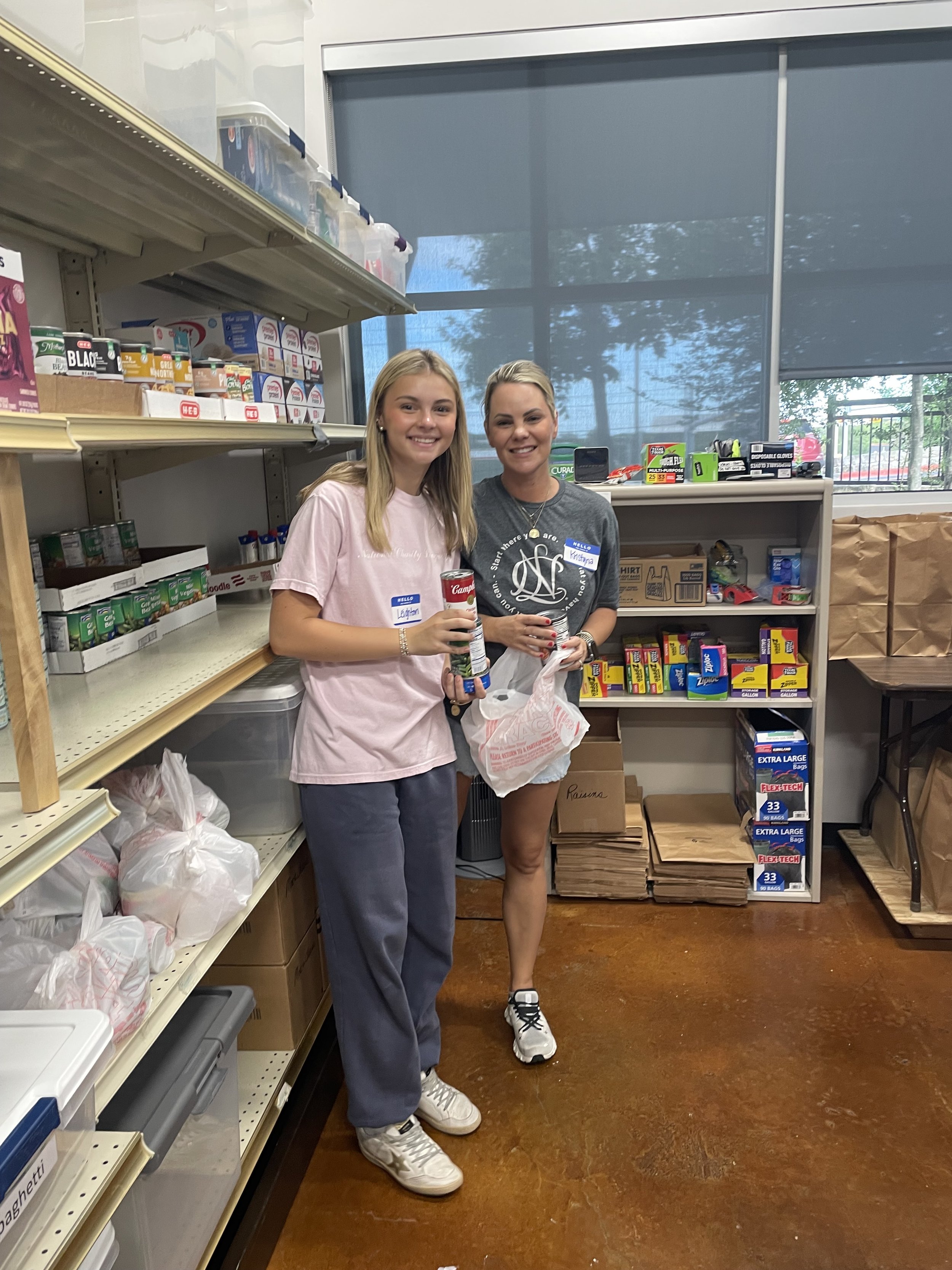 Two women smiling and standing in a grocery store aisle, holding cans and a plastic bag, with shelves stocked with canned goods and household items behind them.