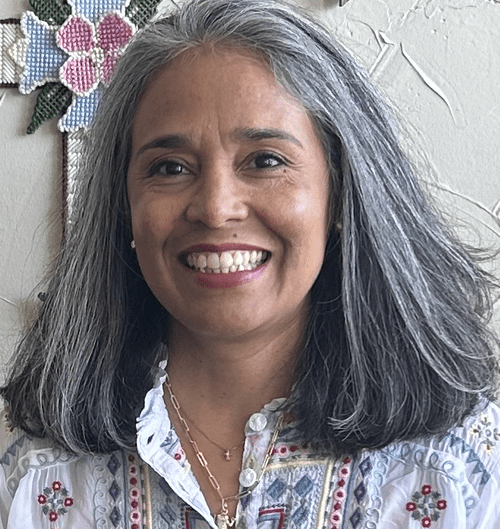 A woman with long gray hair smiling, wearing a white embroidered blouse and necklaces, standing in front of a white wall with colorful beadwork.