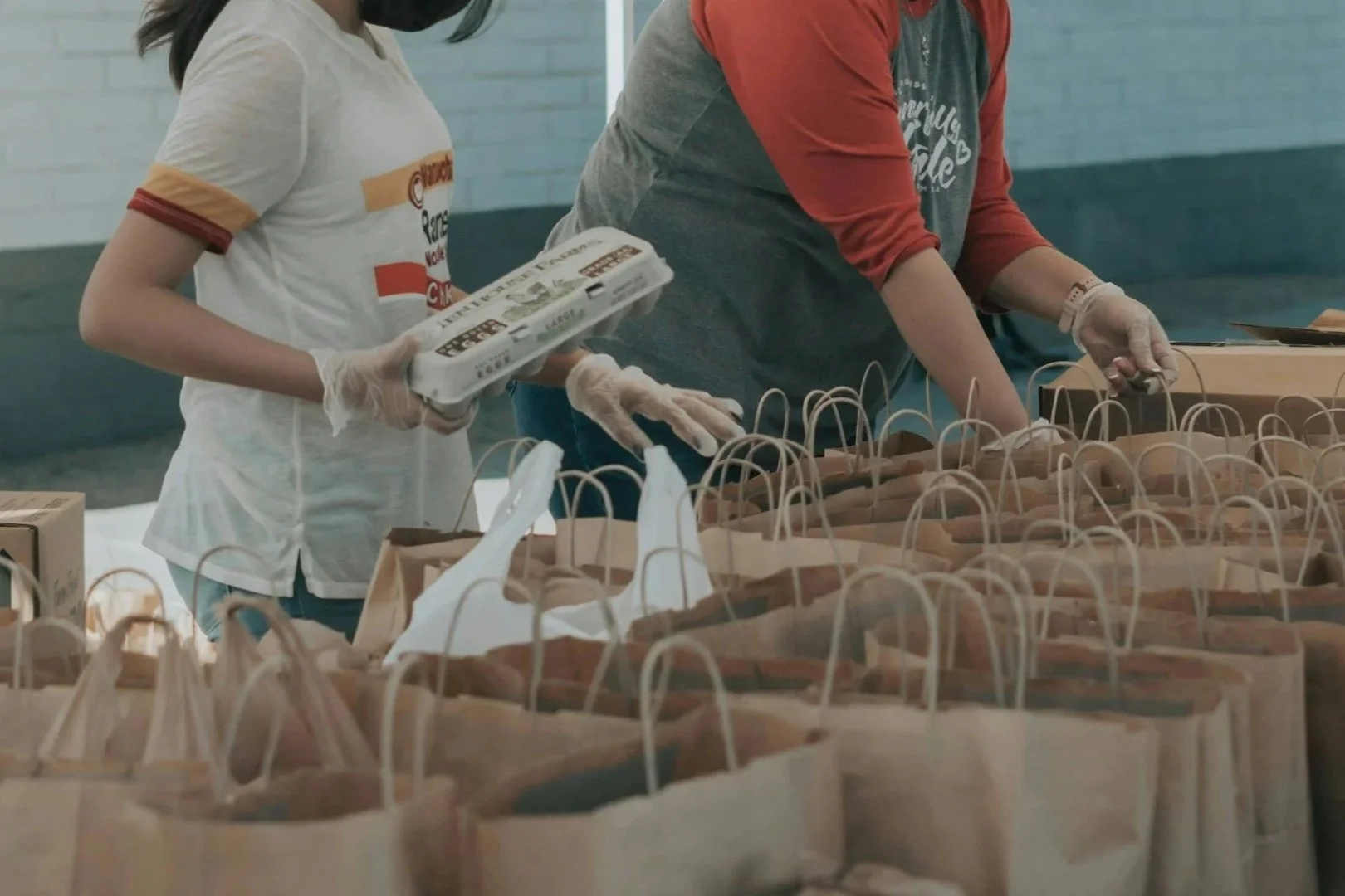 A man wearing a teal shirt, dark blue cap, and gloves is packing or organizing numerous paper grocery bags on a wooden floor in a semi-outdoor setting. There are many more paper bags lined up, and the background shows a screened porch with outdoor scenery including trees.