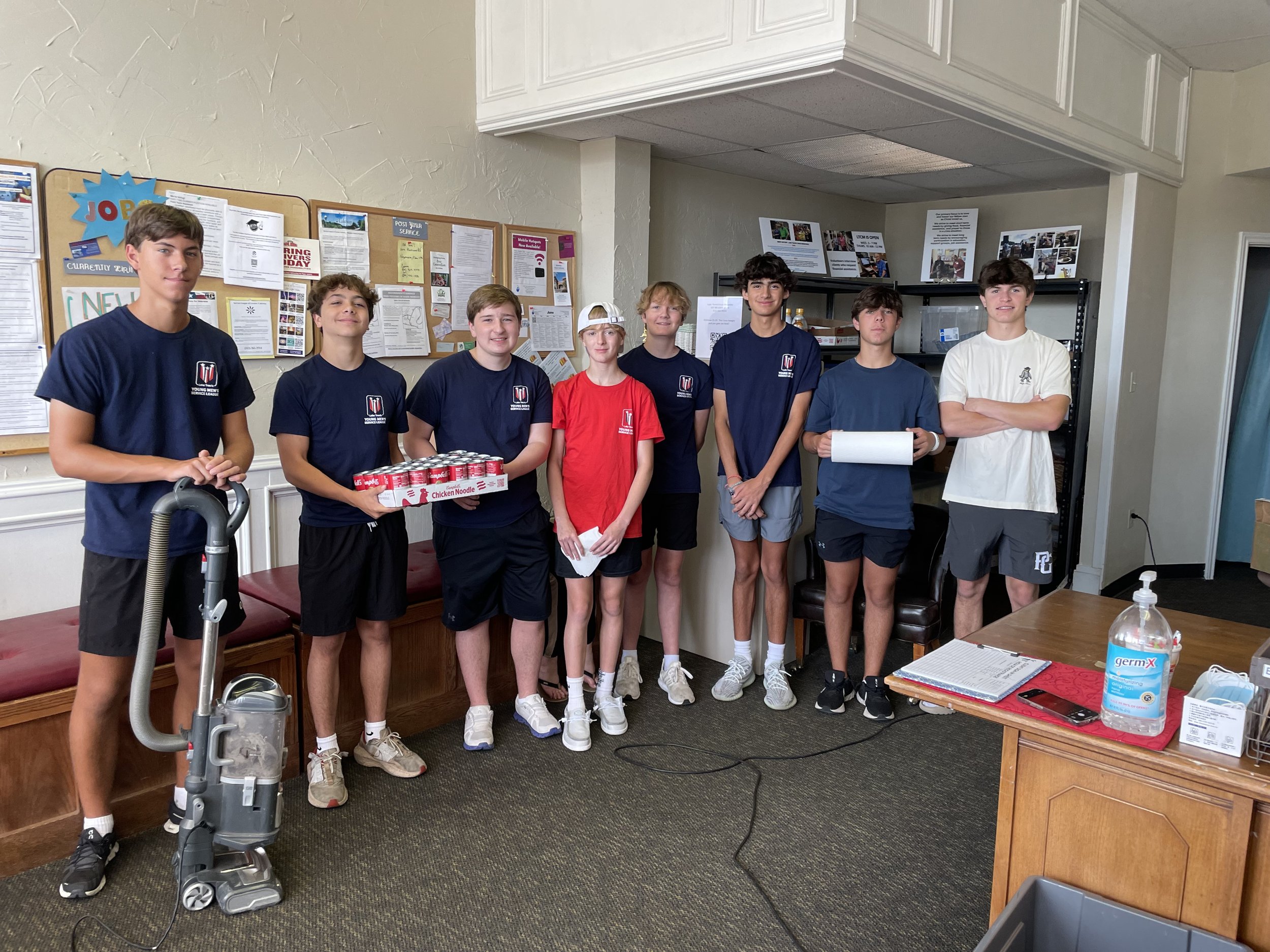 A group of nine young boys standing in a room, some holding boxes of canned food and papers. The room has bulletin boards with notices on the wall behind them and a table with sanitizer and notebooks in front of them.