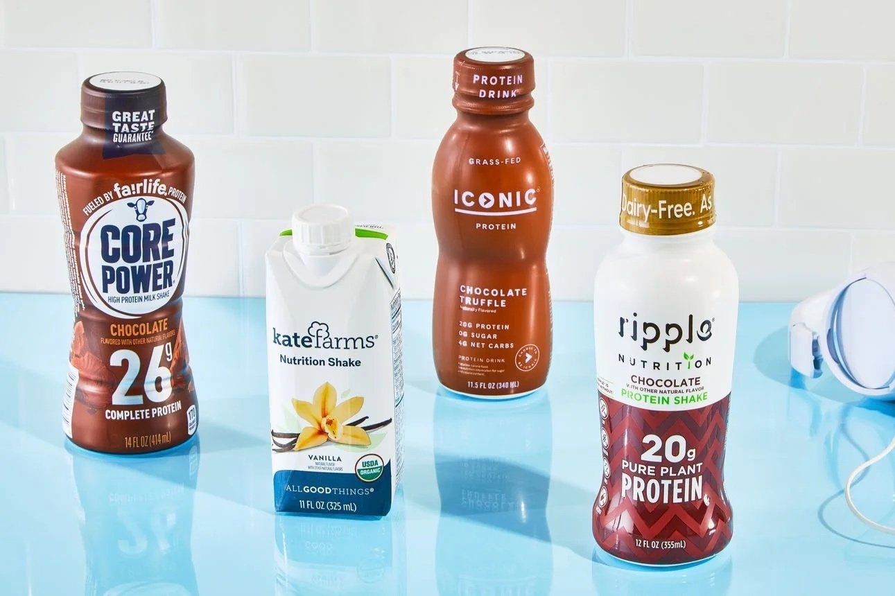 Five protein drink bottles on a blue countertop, with white tiled wall in background. From left to right: Core Power chocolate milk shake, Kate Farms vanilla nutrition shake, Iconic chocolate truffle protein drink, Ripple chocolate protein shake, and a pair of white headphones on the right.