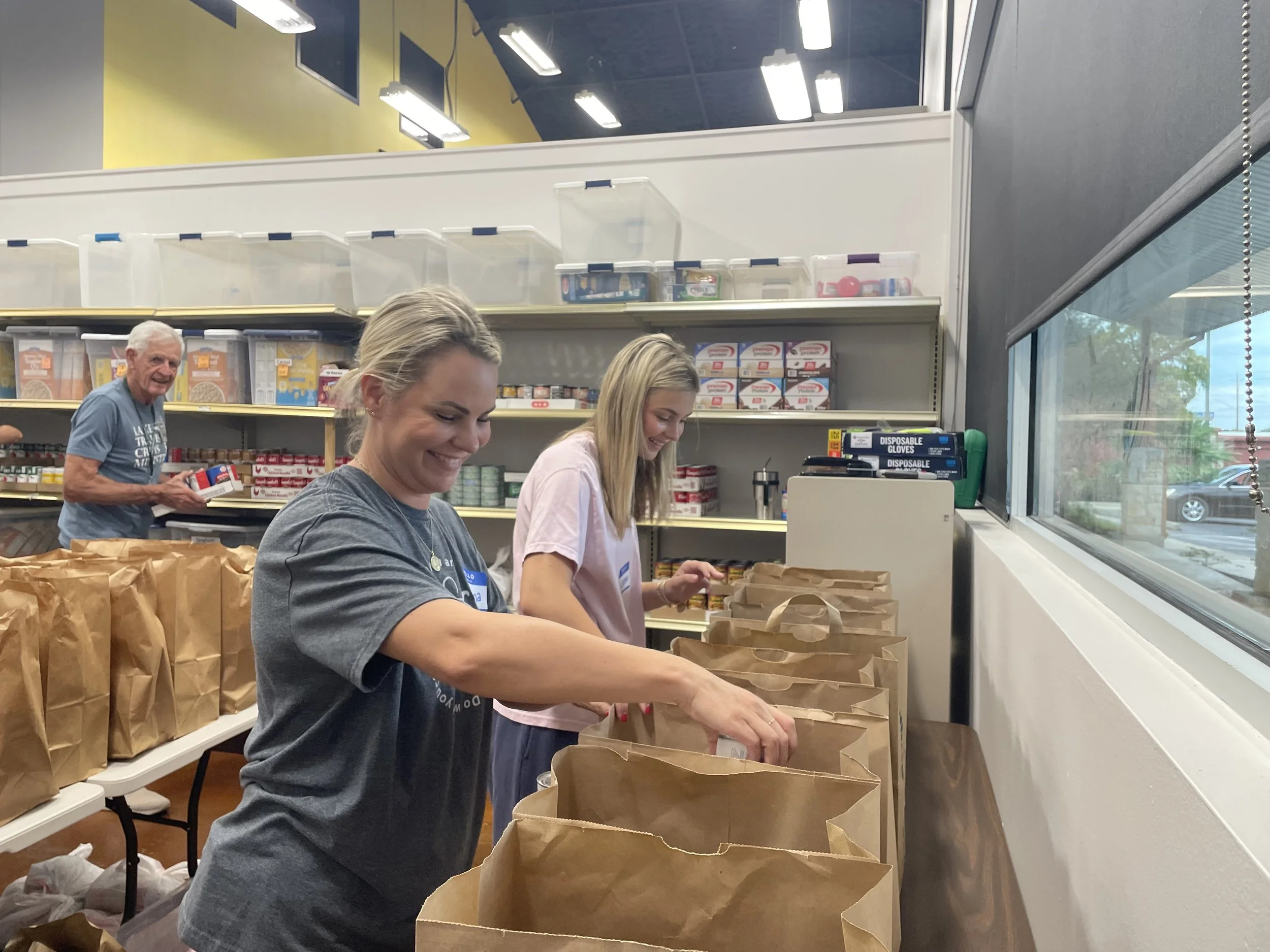 Three women packing brown paper bags at a table inside a store, with shelves of products in the background and a large window on the right.