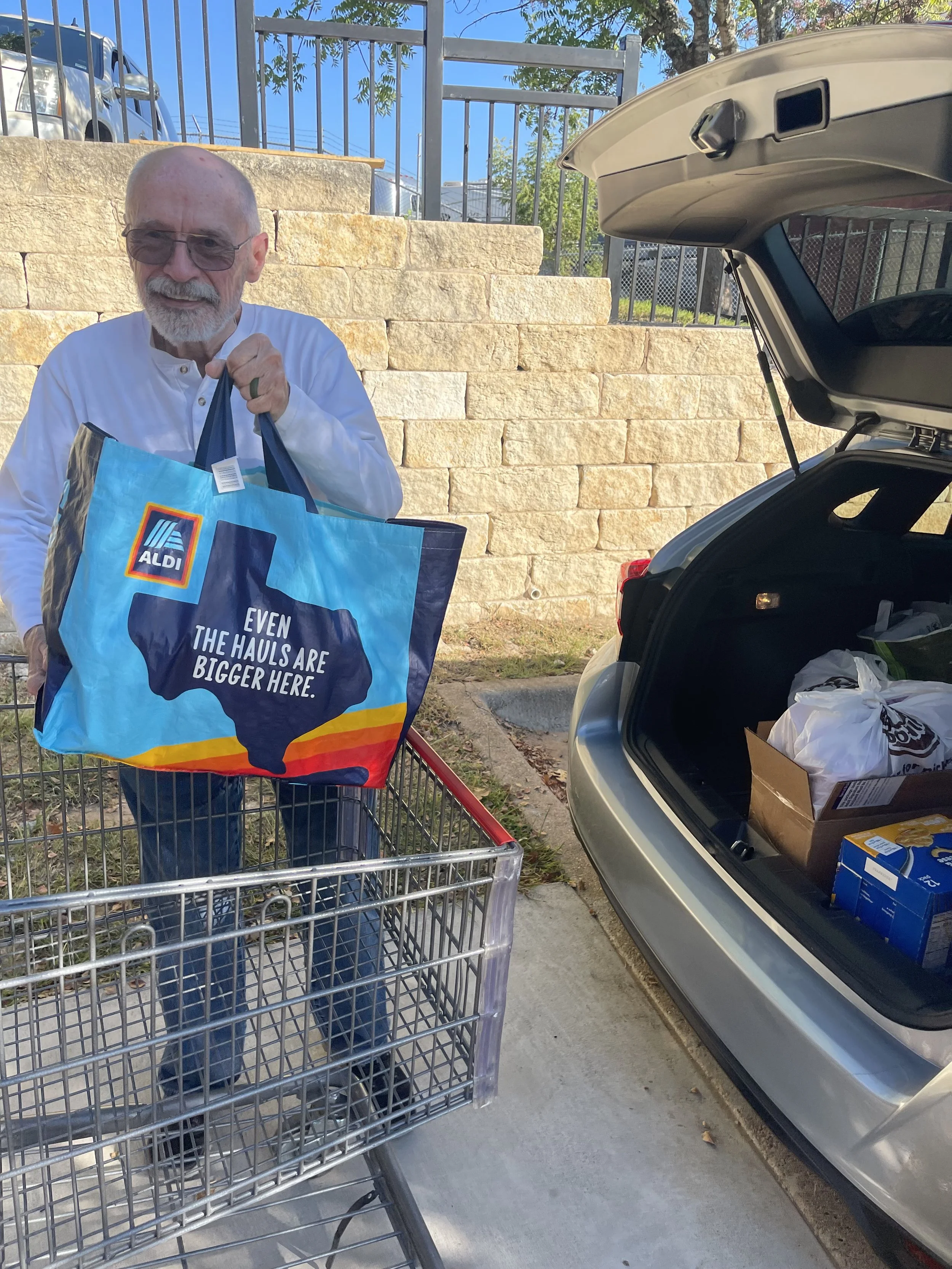 An elderly man with glasses and a white shirt is holding an Aldi shopping bag with a Texas outline and the text 'Even the haulers are bigger here.' He is standing next to a shopping cart and near an open trunk of a car filled with grocery bags, in a parking lot with a stone wall and metal fence in the background.