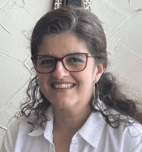 A woman with curly brown hair, wearing red glasses and a white shirt, smiling at the camera.