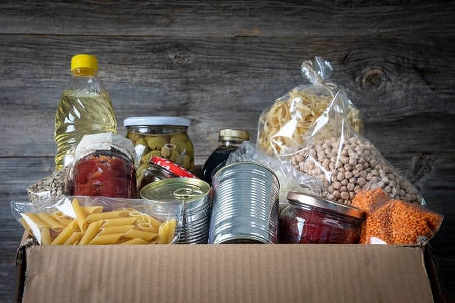 A cardboard box filled with various canned and packaged food items, including pasta, canned vegetables, jars, a bottle of cooking oil, and dried lentils, set against a wooden background.