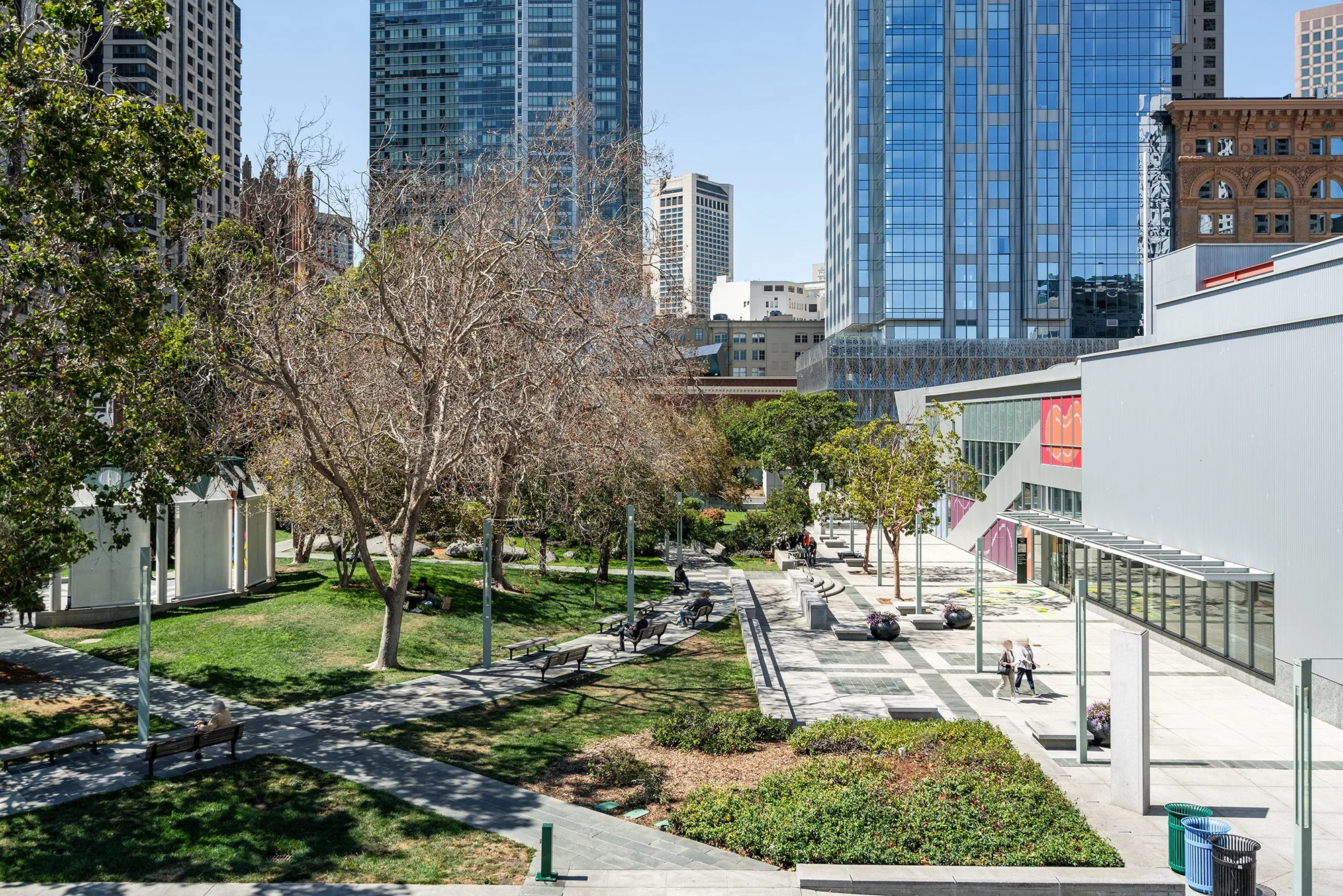 Urban park with trees, benches, and people walking on paved pathways surrounded by tall modern city buildings.