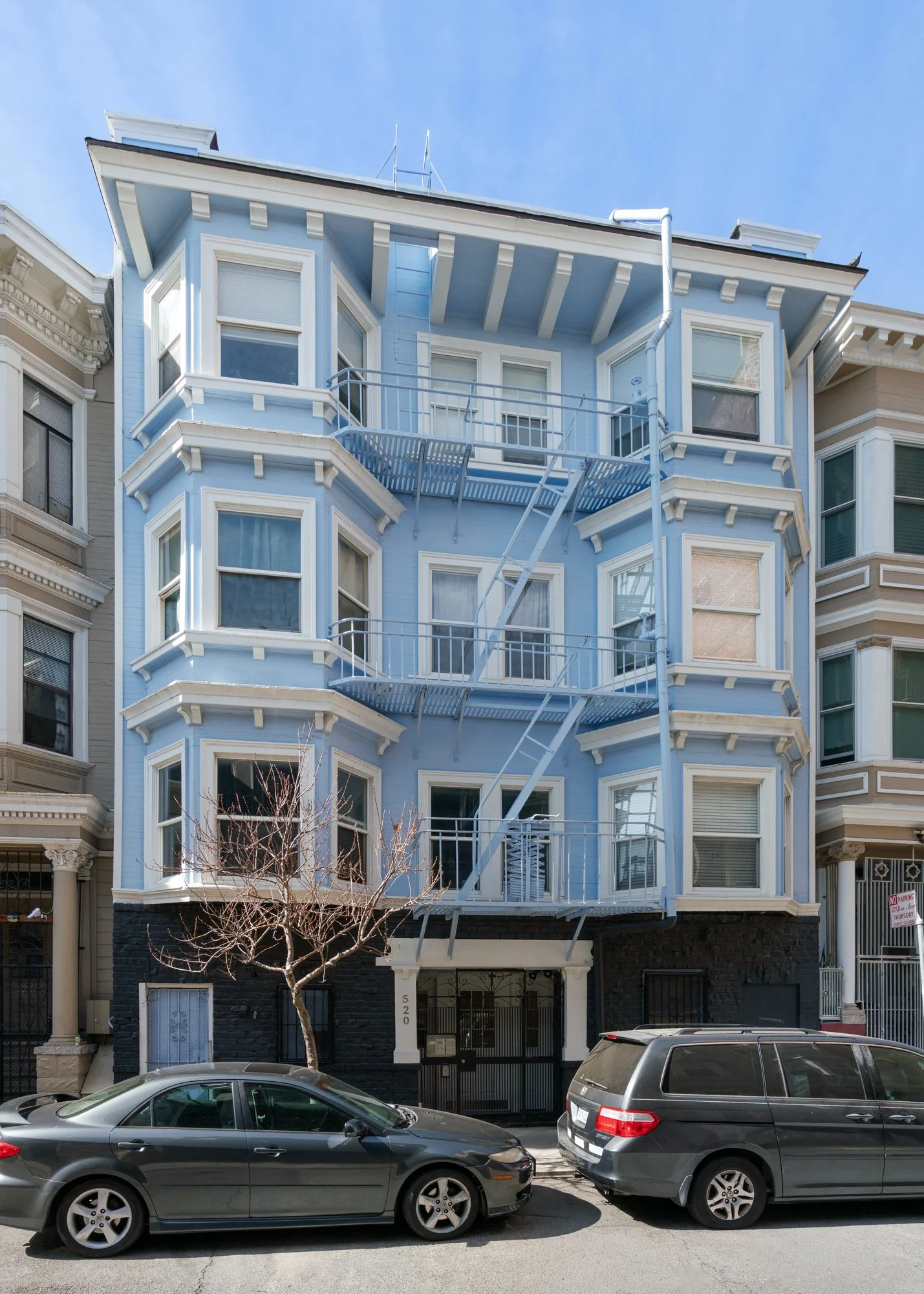 Blue multi-story residential building with fire escape staircases on the front, parked cars at street level, and a leafless tree in front.