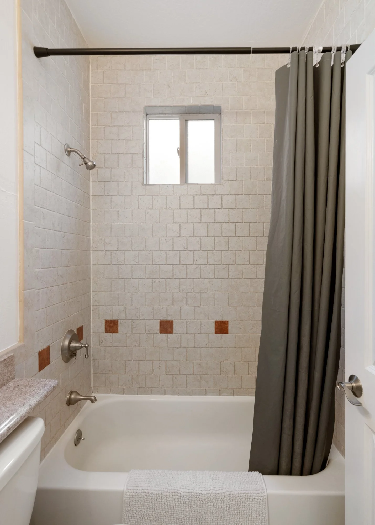 Bathroom with bathtub, dark gray shower curtain, small window, beige tiled walls, and beige tiles with orange accent tiles.