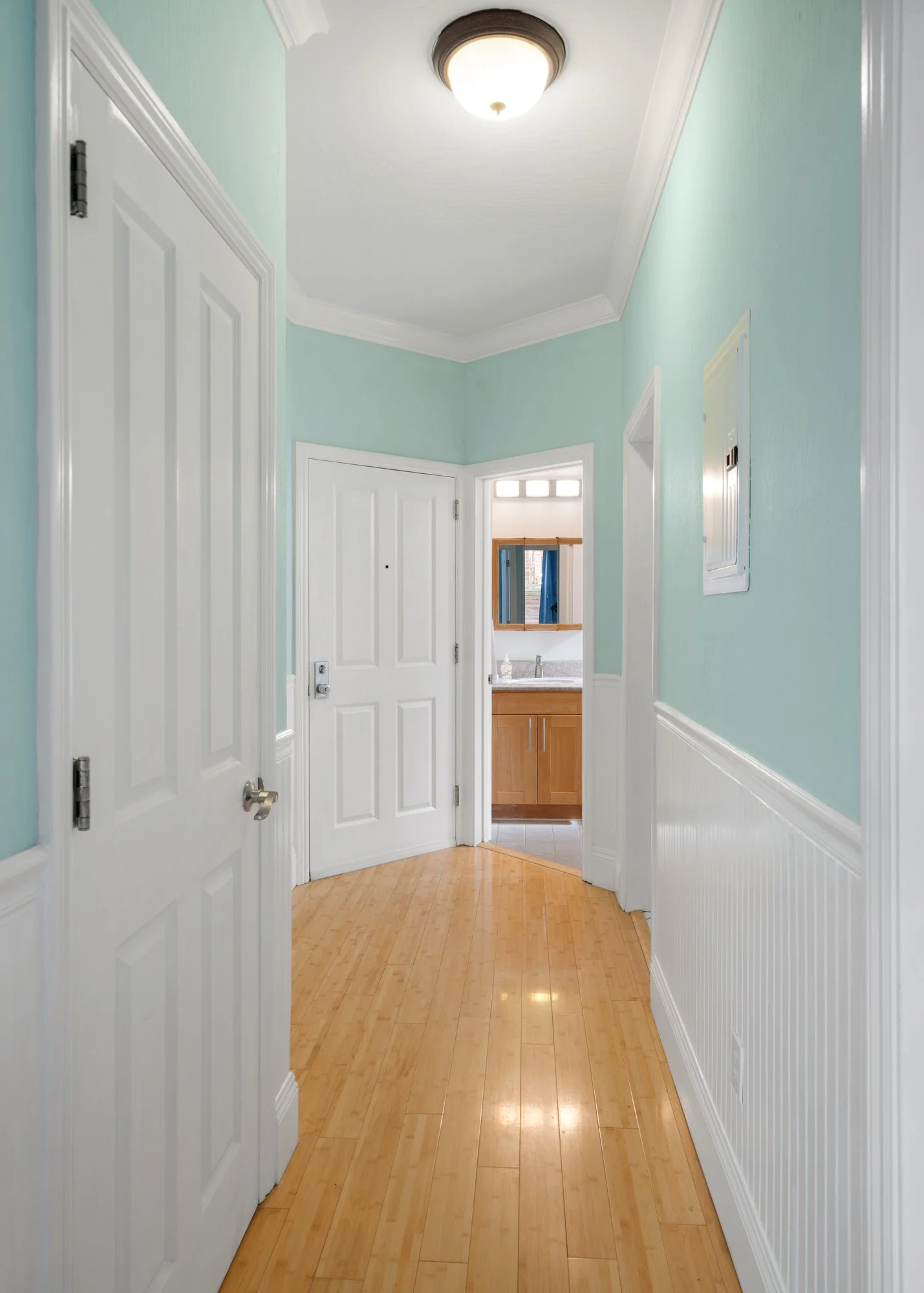 Empty hallway with light blue walls, white trim, and wooden floors leading to a bathroom with wooden cabinetry and a mirror.