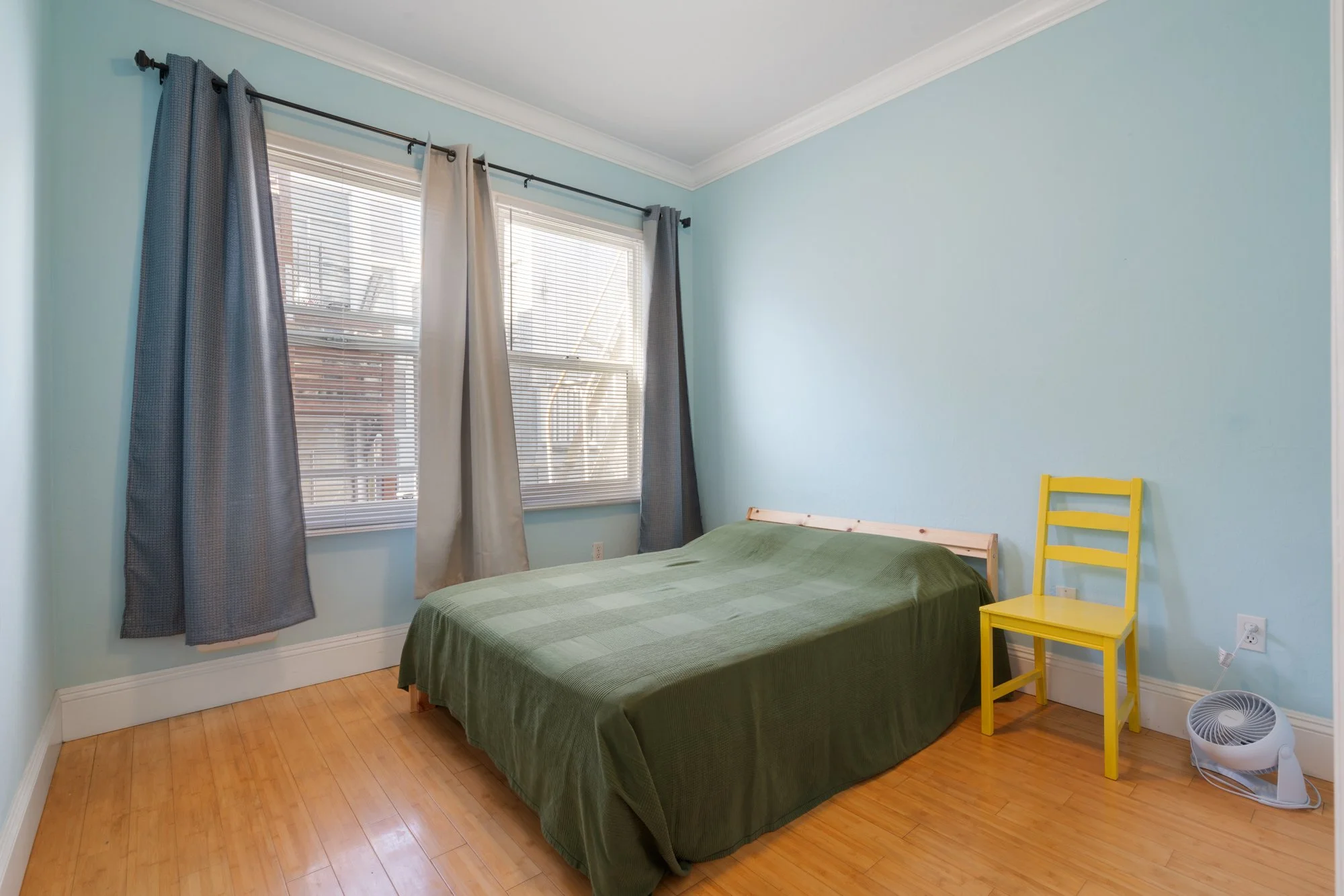 Empty bedroom with light blue walls, hardwood floor, and two windows with blinds and curtains. Contains a bed with a green blanket, a yellow chair, and a small portable fan on the floor.