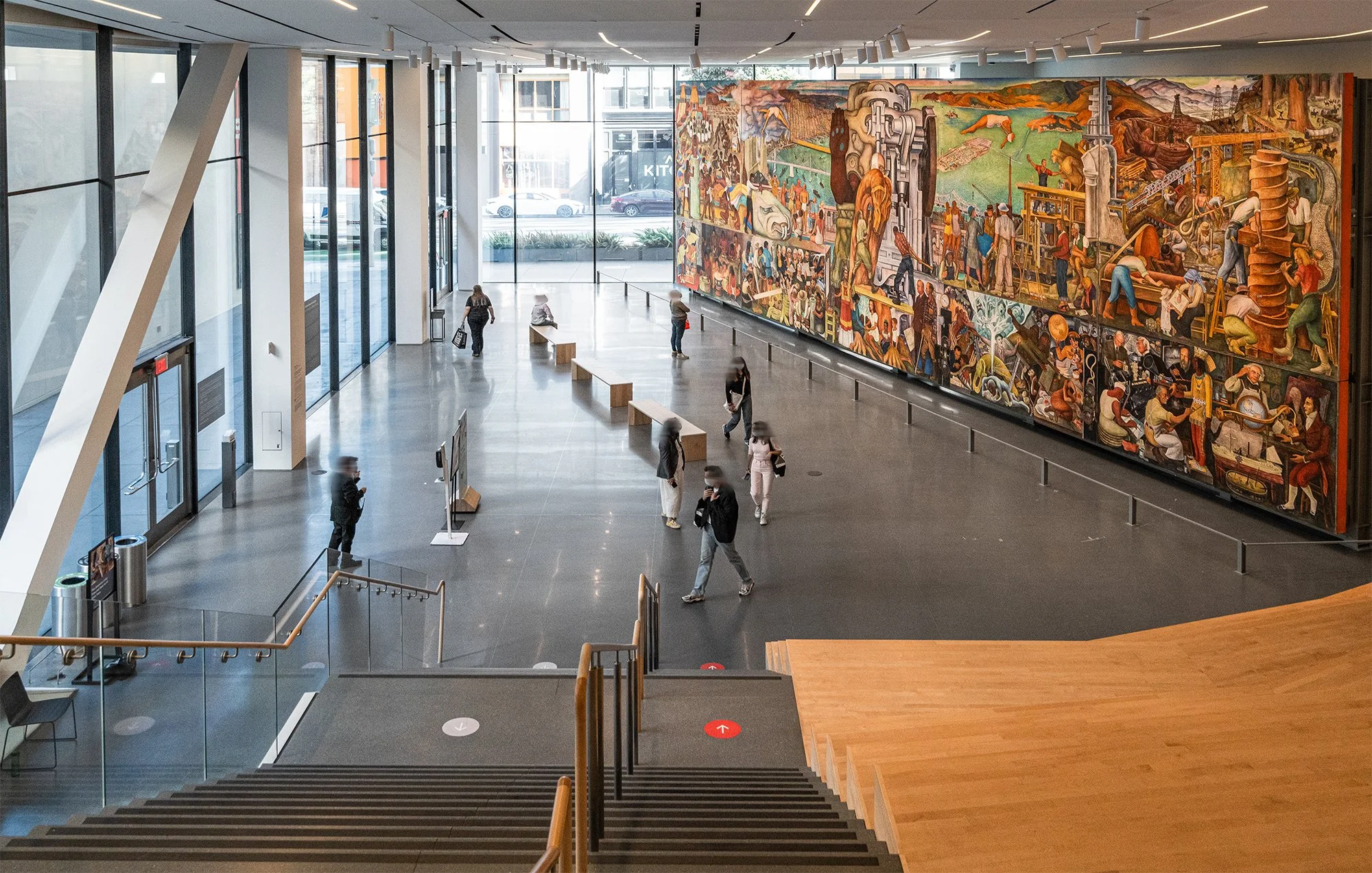 Interior of a modern building with large glass windows, a colorful mural on the wall, and people walking or sitting in the spacious lobby area.