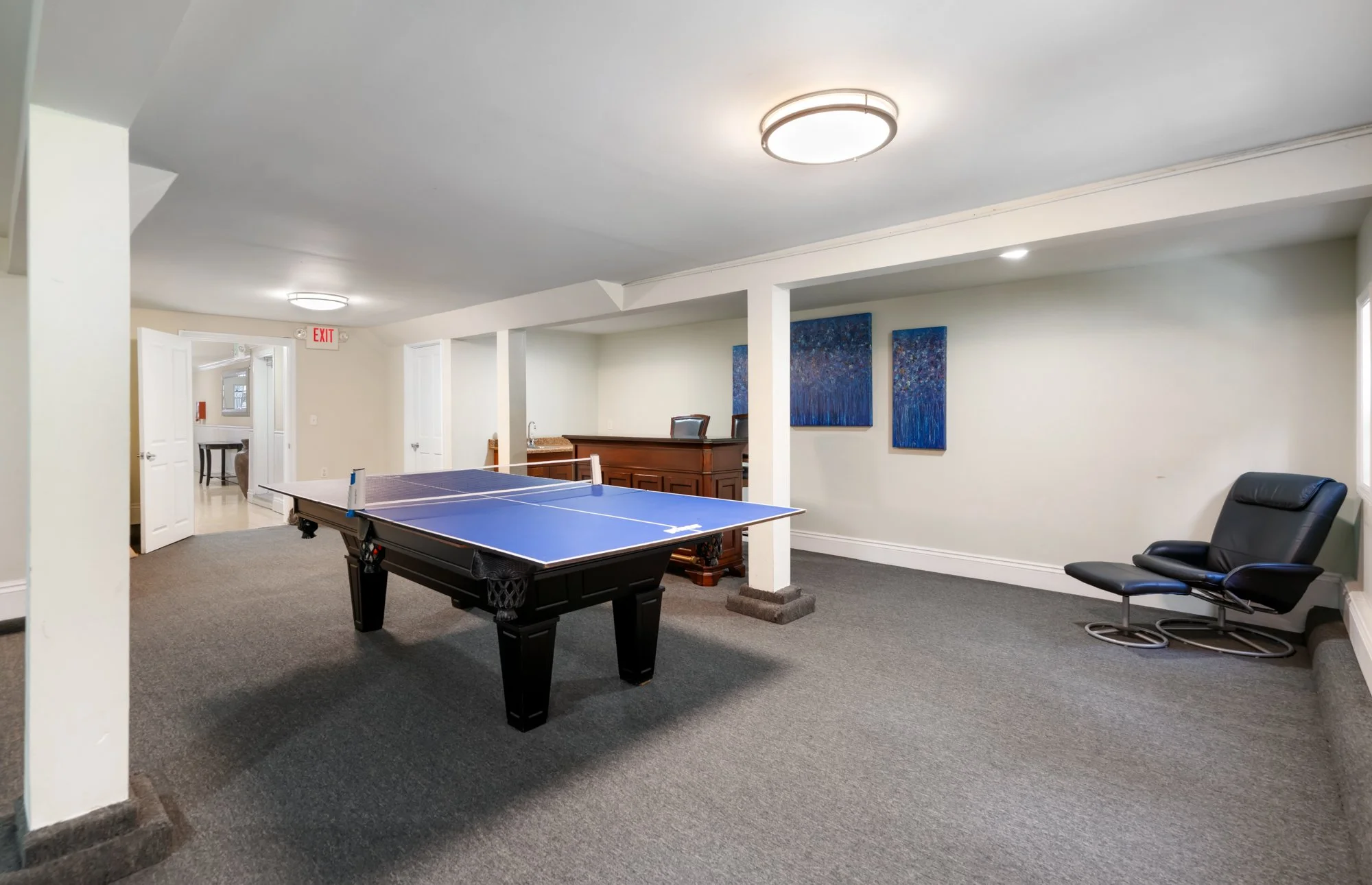 A basement recreation room with a ping pong table, a black lounge chair, blue artwork on the wall, and a wooden bar with a chair.