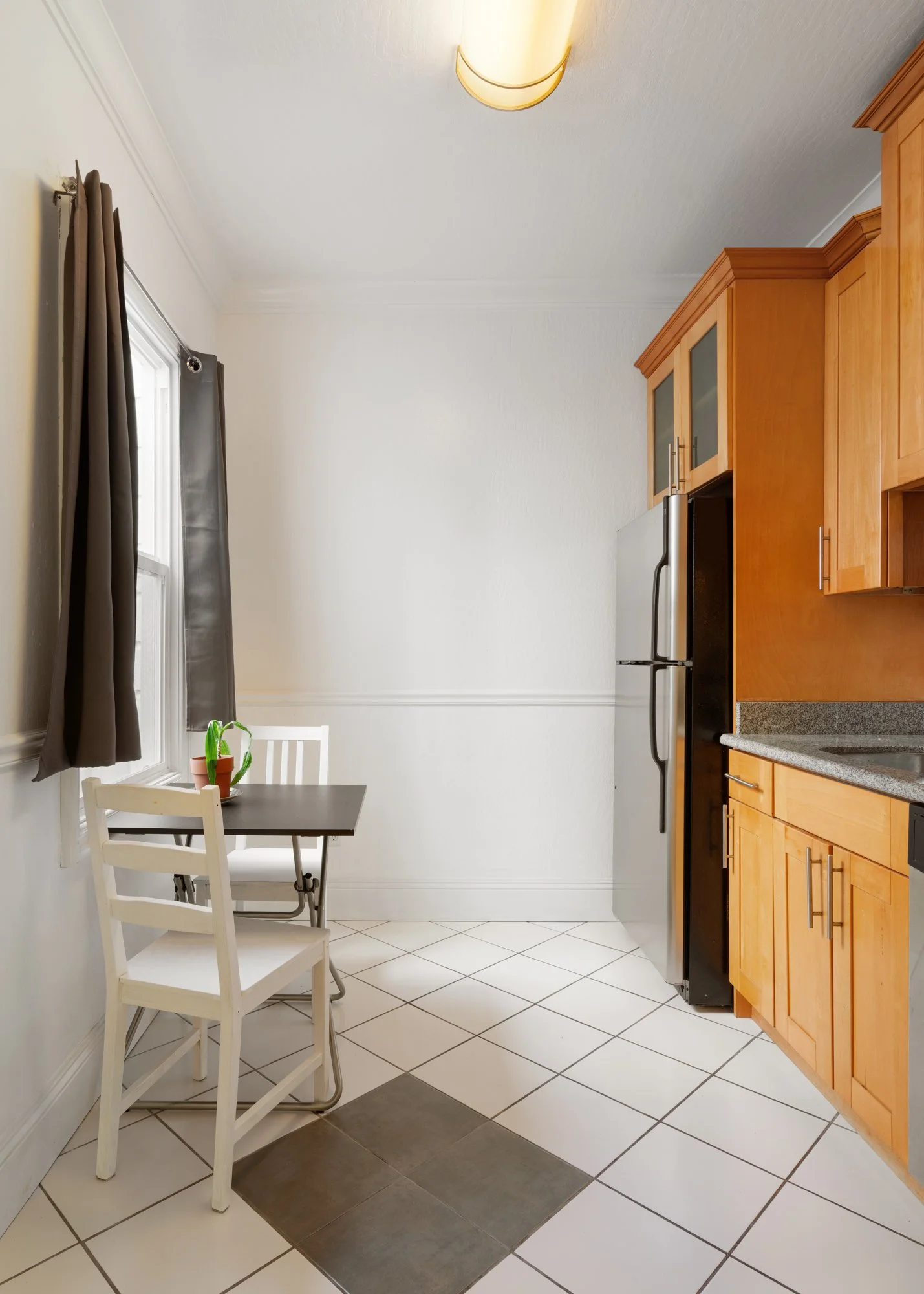 Kitchen corner with a small dining table and two white chairs by a window, wooden cabinets, a refrigerator, and tiled floor.