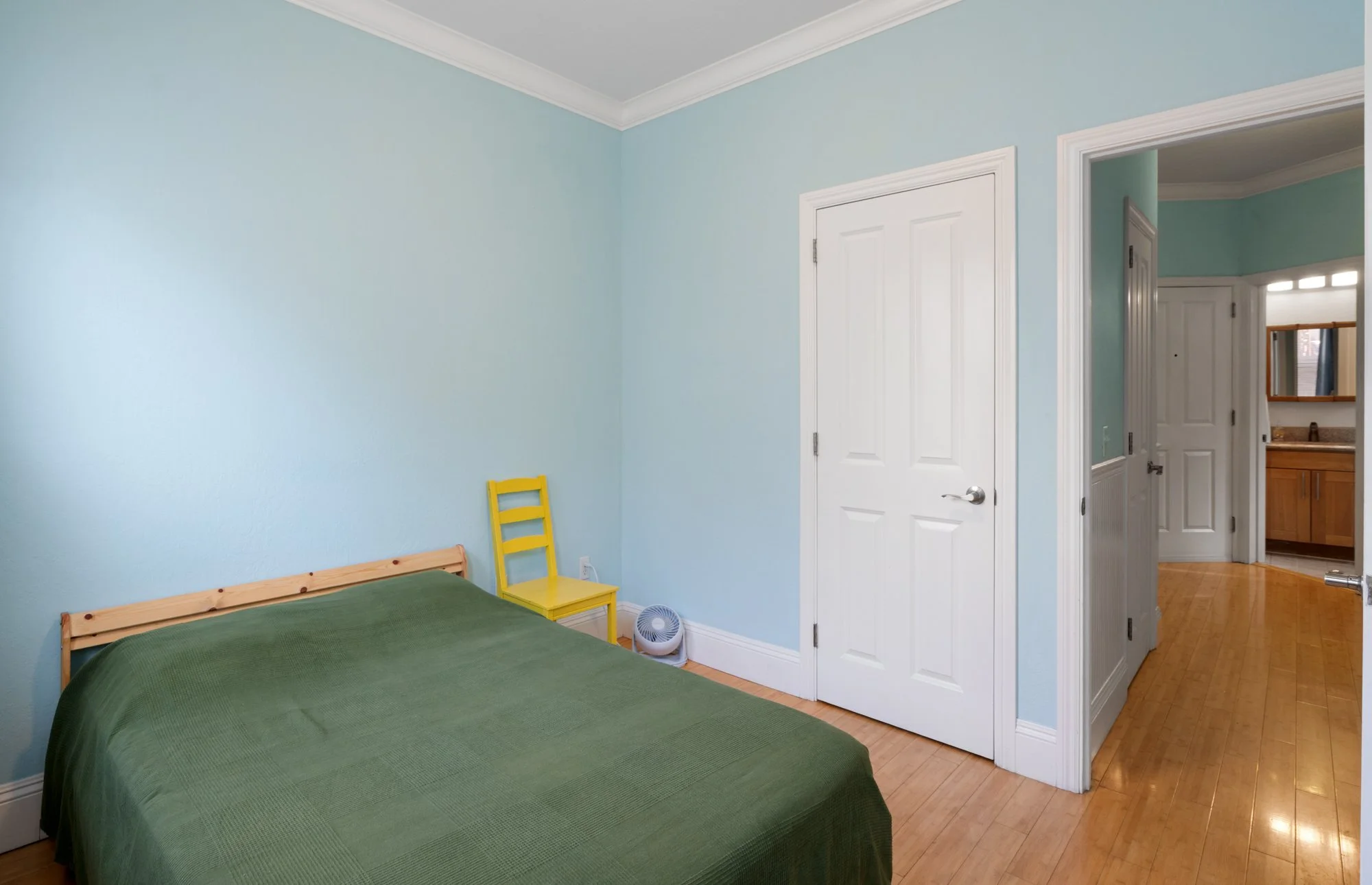 A bedroom with light blue walls, a bed with a green cover, a yellow chair, a small fan, and a doorway leading to a hallway and bathroom.