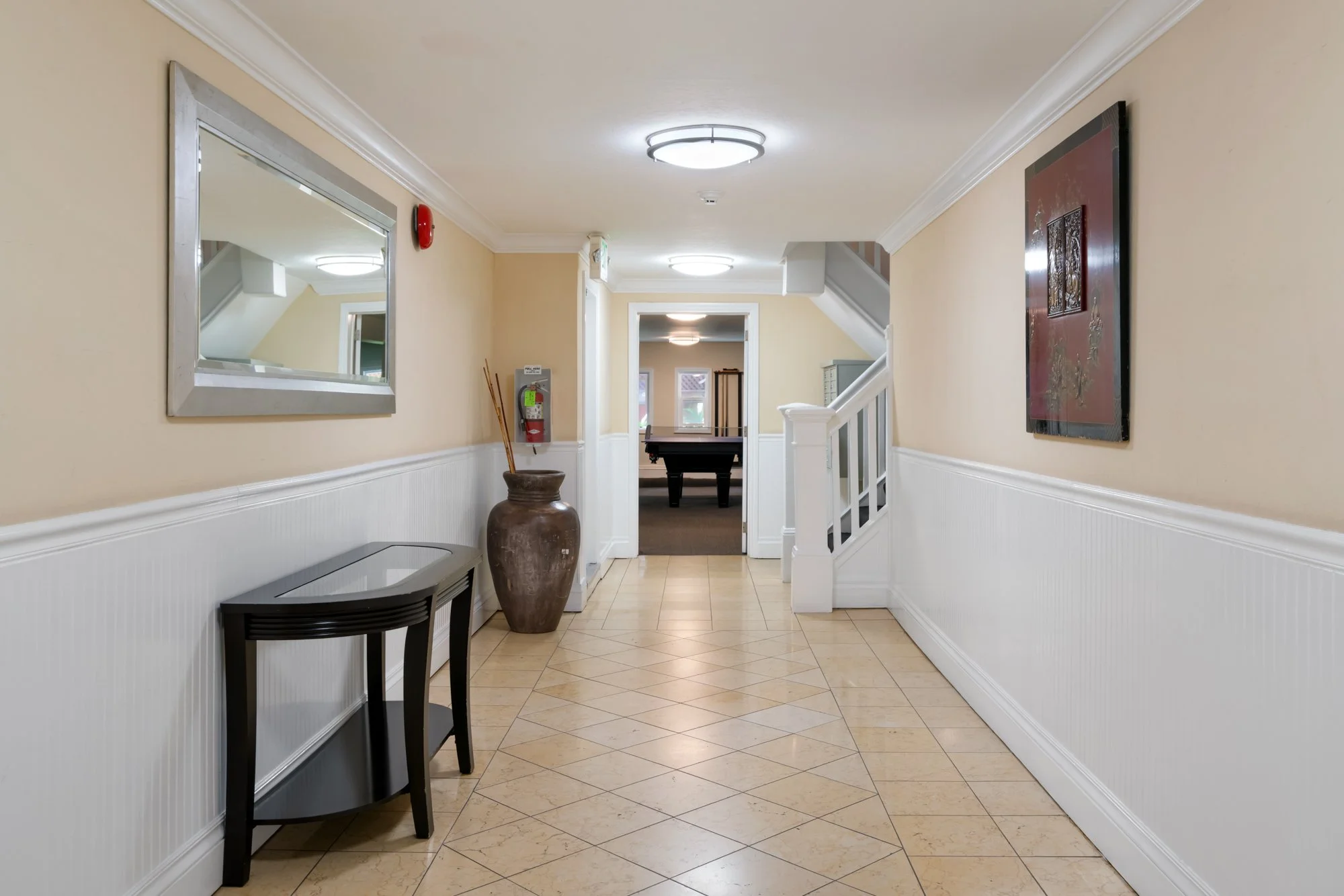 Indoor hallway with beige walls, white wainscoting, tiled floor, large wall mirror, wall-mounted fire alarm, fire extinguisher, decorative vase with sticks, staircase, and artwork, leading to a room with a pool table.