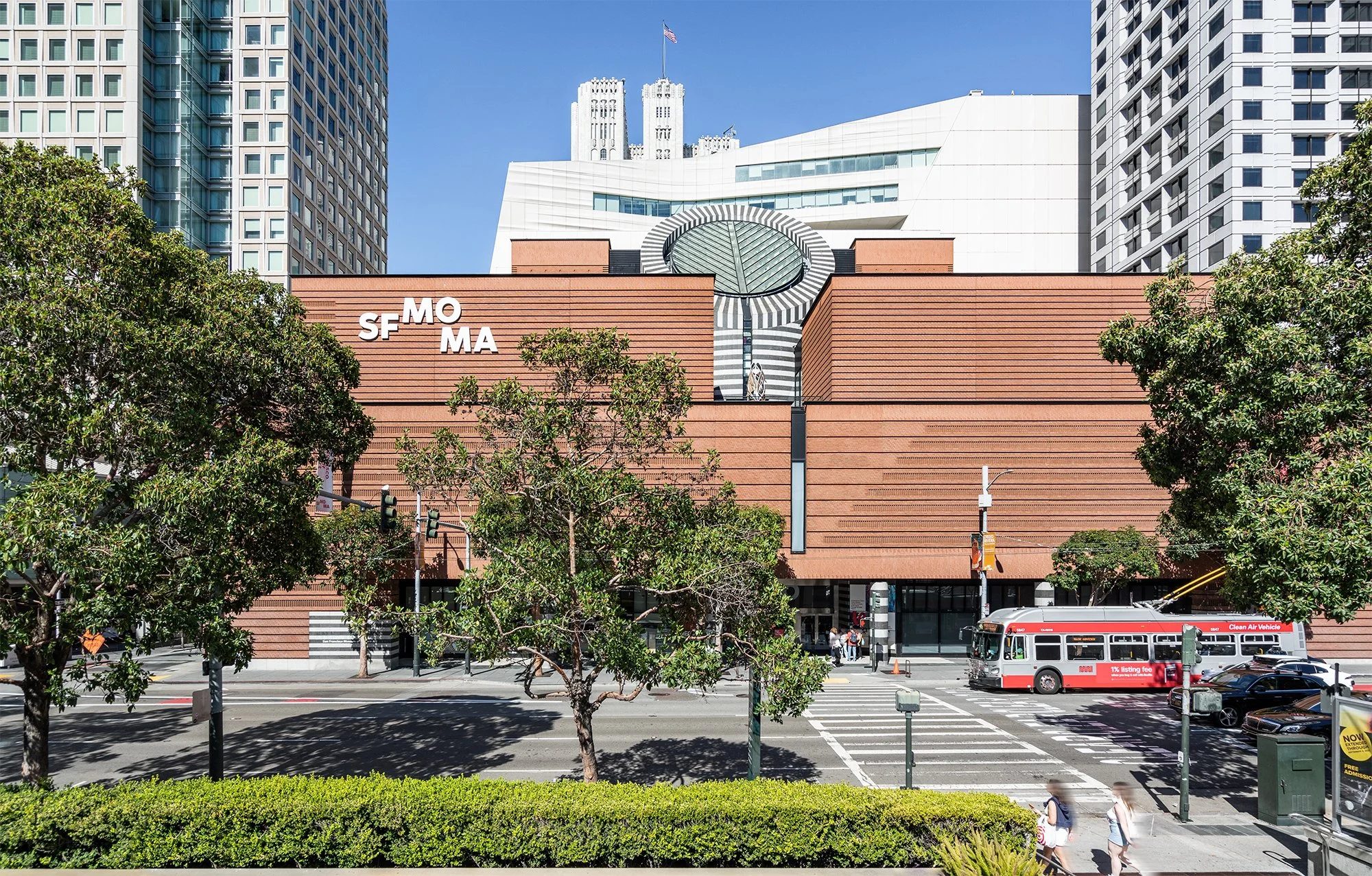 Exterior view of the San Francisco Museum of Modern Art (SFMOMA) with the museum's entrance, trees, a bus, crosswalk, and pedestrians in the foreground, and taller buildings behind it under a clear blue sky.
