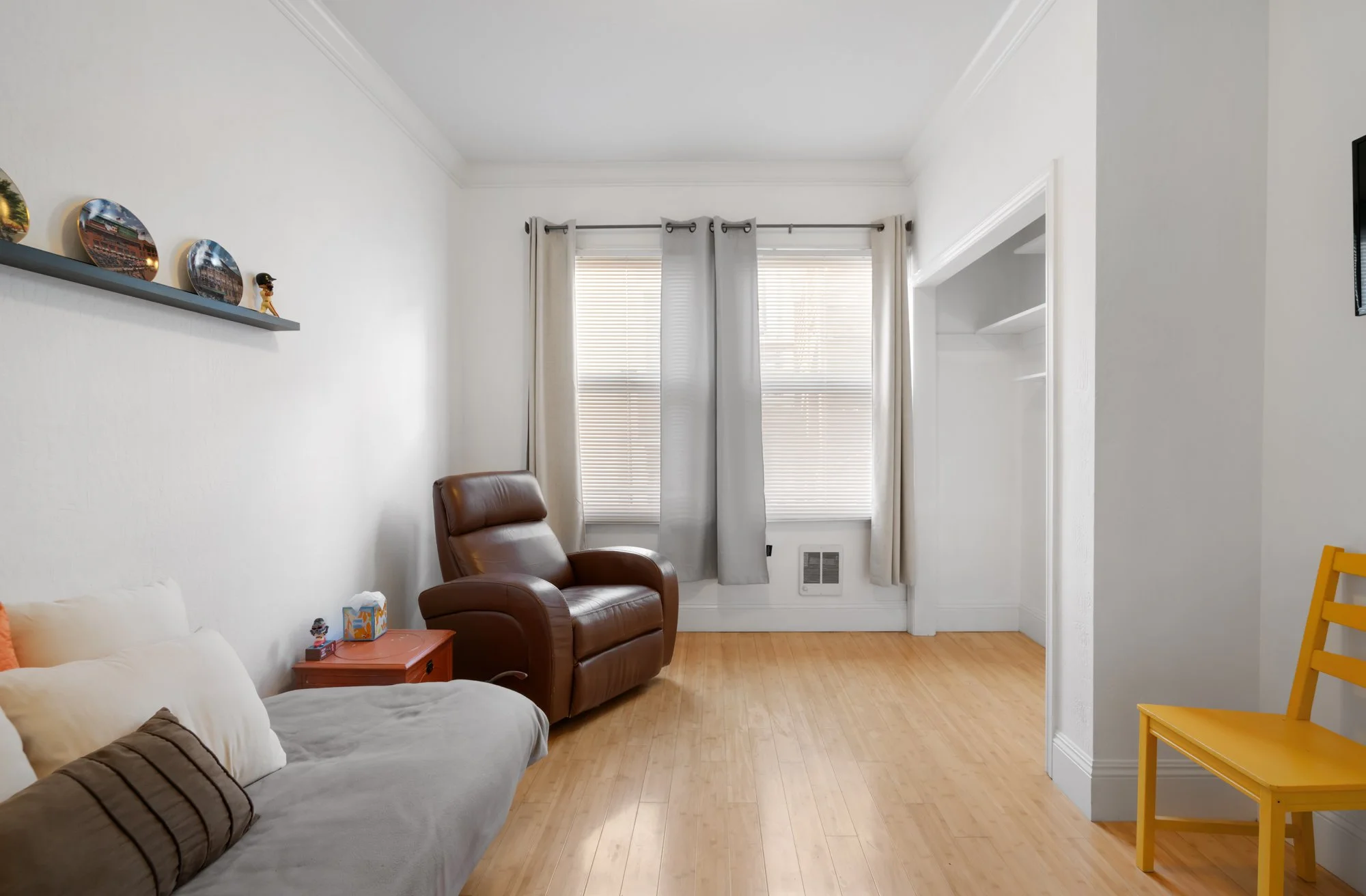 A bright living room with light wood flooring, white walls, and two large windows with blinds and beige curtains. There is a beige sofa with cushions on the left, a brown leather armchair near the window, a small red side table with a tissue box and 