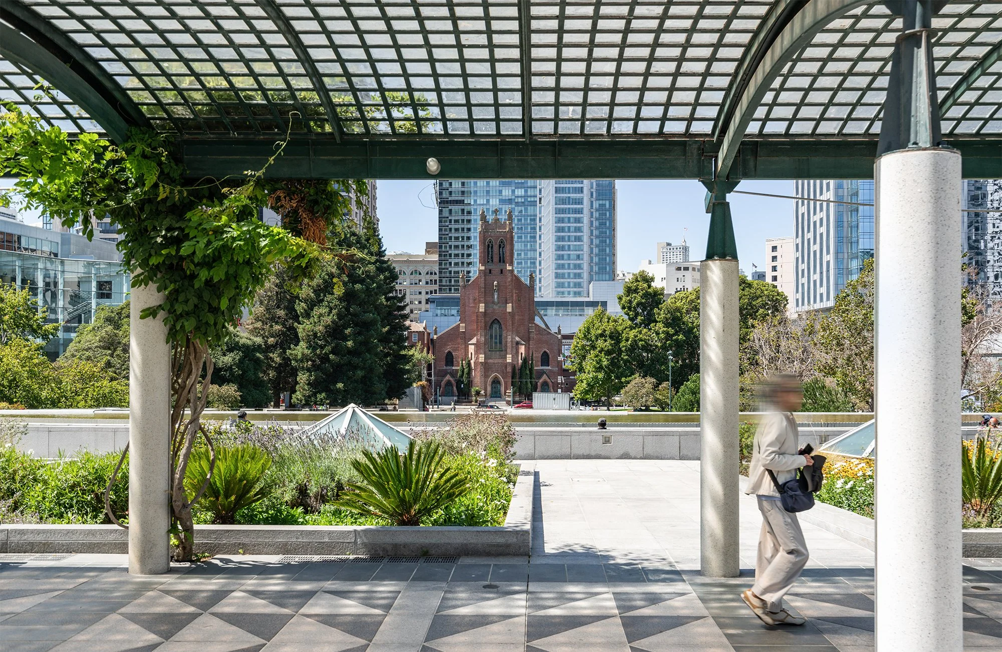 Cityscape view from under a covered walkway, with a person walking by in the foreground, and a red brick church surrounded by green trees and modern skyscrapers in the background.