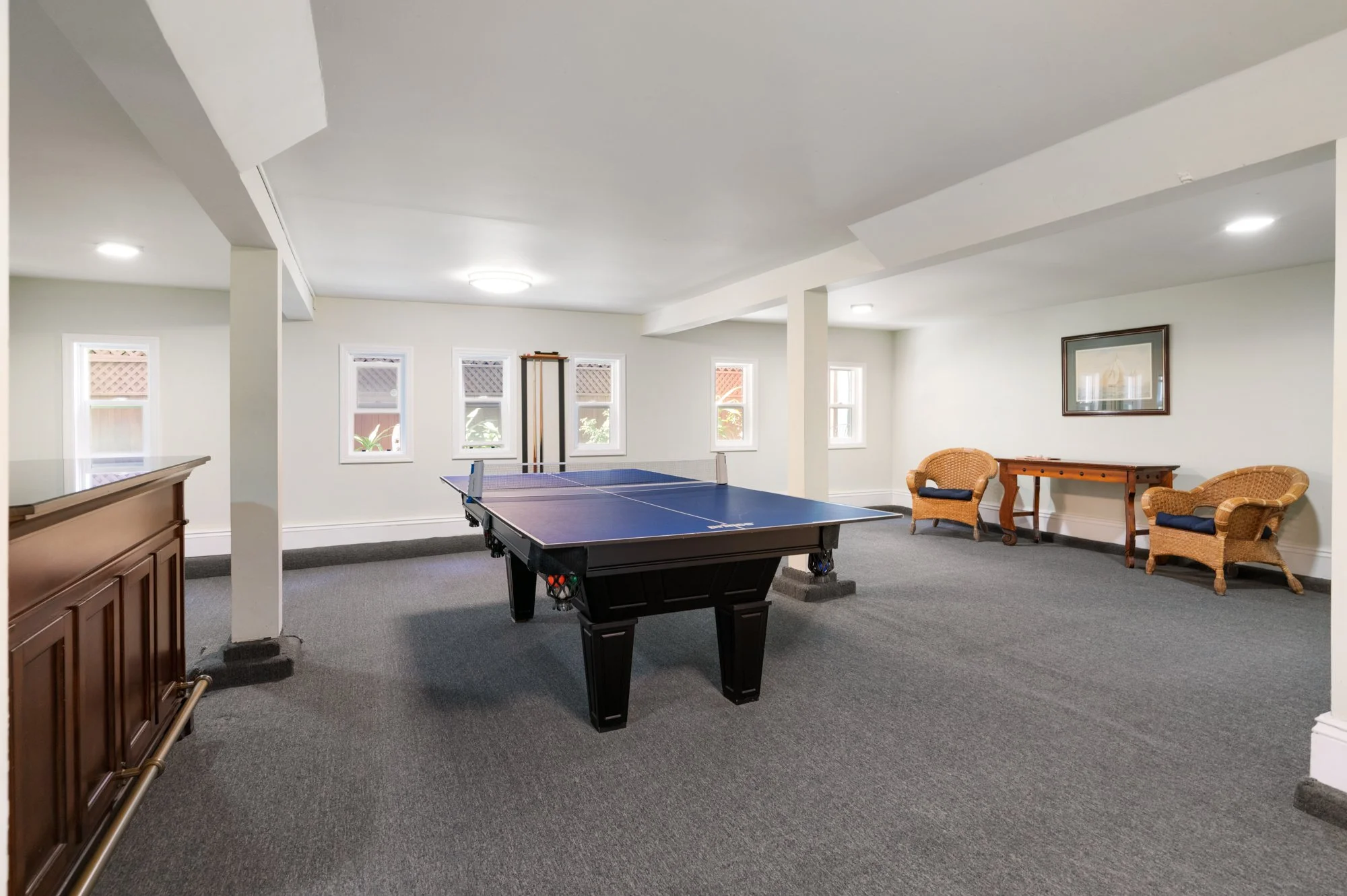 A game room with a ping pong table, two wicker chairs, a small wooden table, and a framed picture on the wall, with four windows letting in natural light.