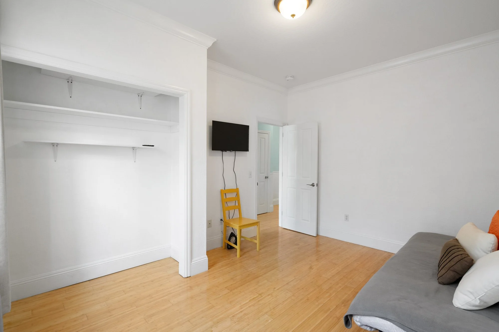 Empty room with white walls and ceiling, light wood floor, open closet with no doors, a mounted TV with visible cords, a yellow chair, and part of a gray sofa with cushions.