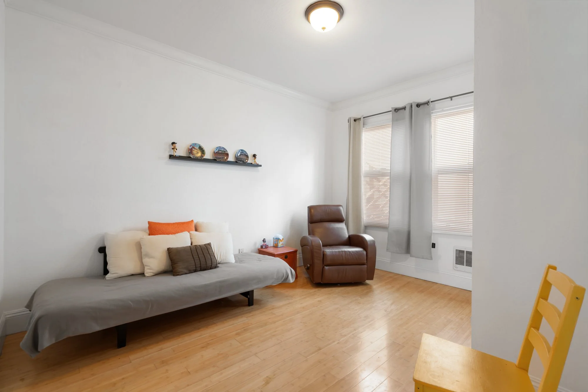Living room with a white wall, a brown leather armchair, a small red side table, a dark gray futon with multiple pillows, and a large window with blinds and curtains.